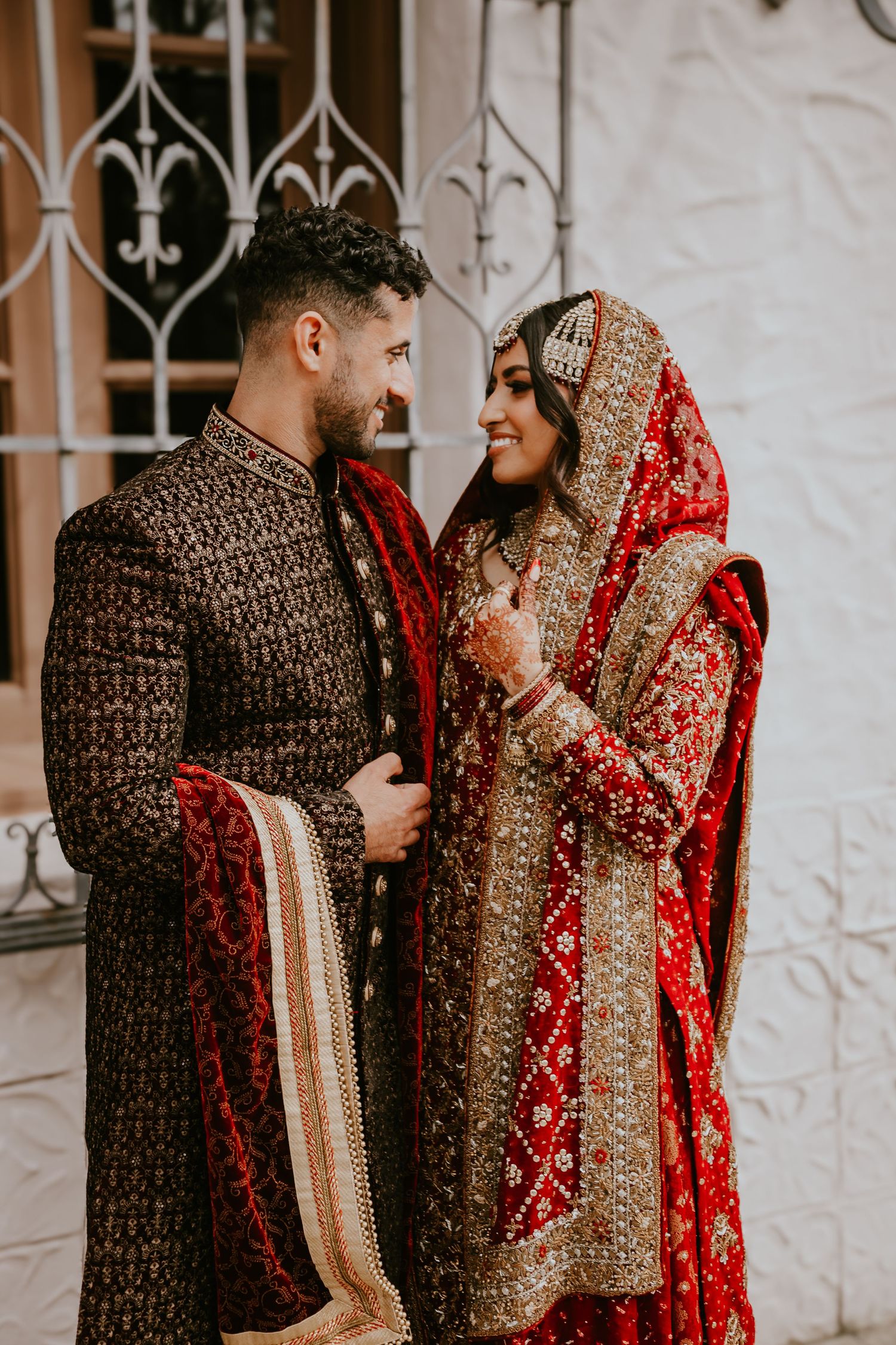 Traditional South Asian couple share a joyful moment wearing ornate wedding attire with red and gold embroidered details.