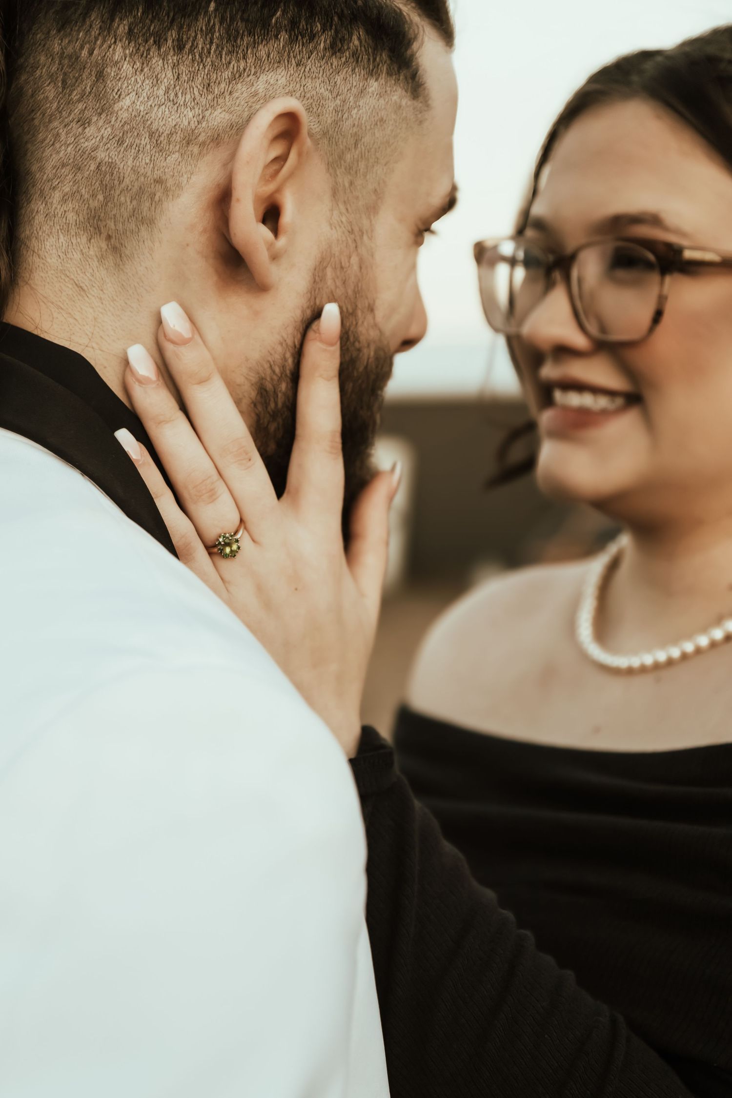 An intimate wedding moment shows a tender touch between partners wearing white and black formal attire with a pearl necklace detail.