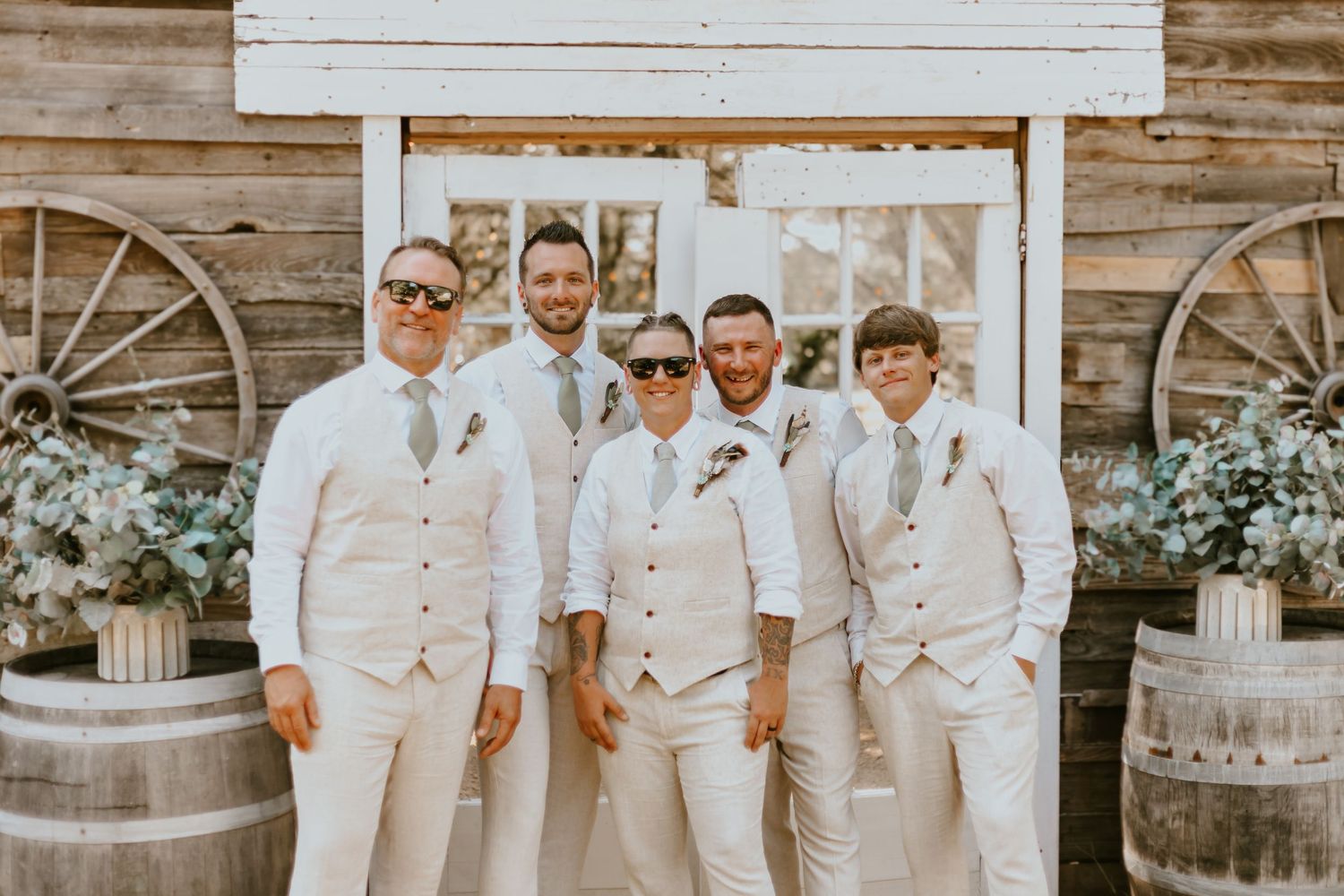 Wedding party members in light beige and white attire pose together in front of a rustic wooden barn backdrop.