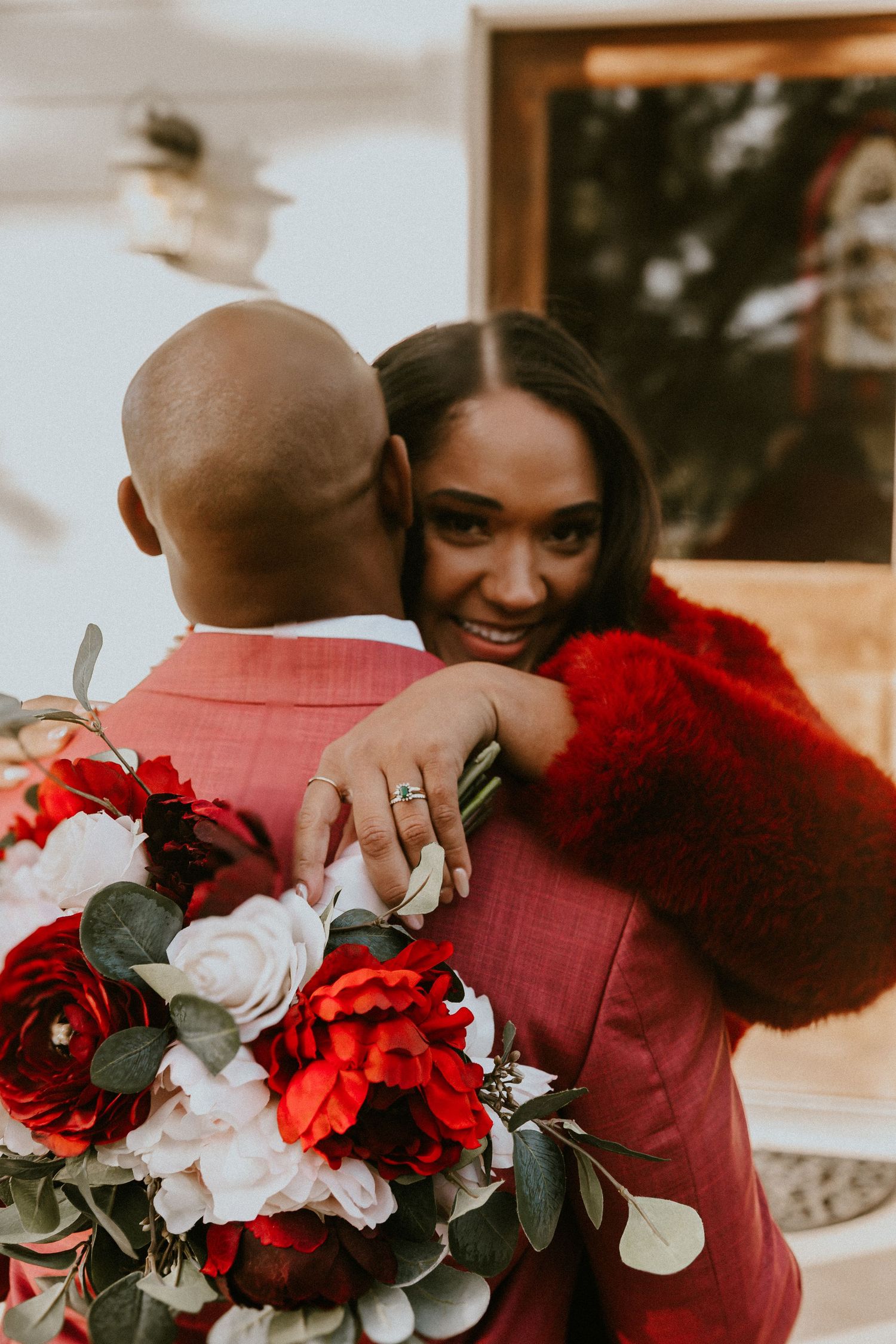A romantic couple embraces while holding a stunning bouquet of red and white flowers against a warm outdoor setting.