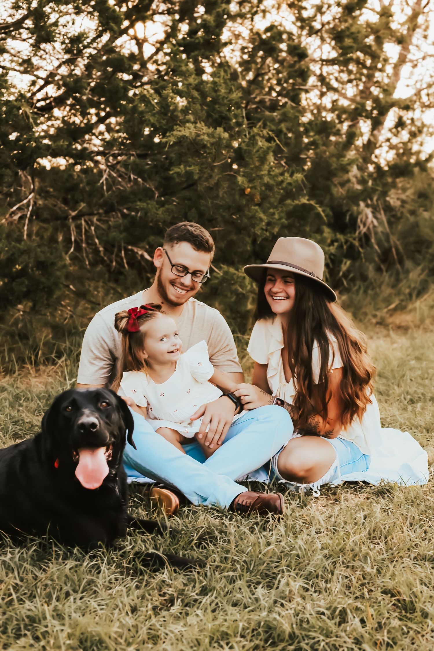 A family and their black dog sit together in a grassy field at sunset with trees in the background.