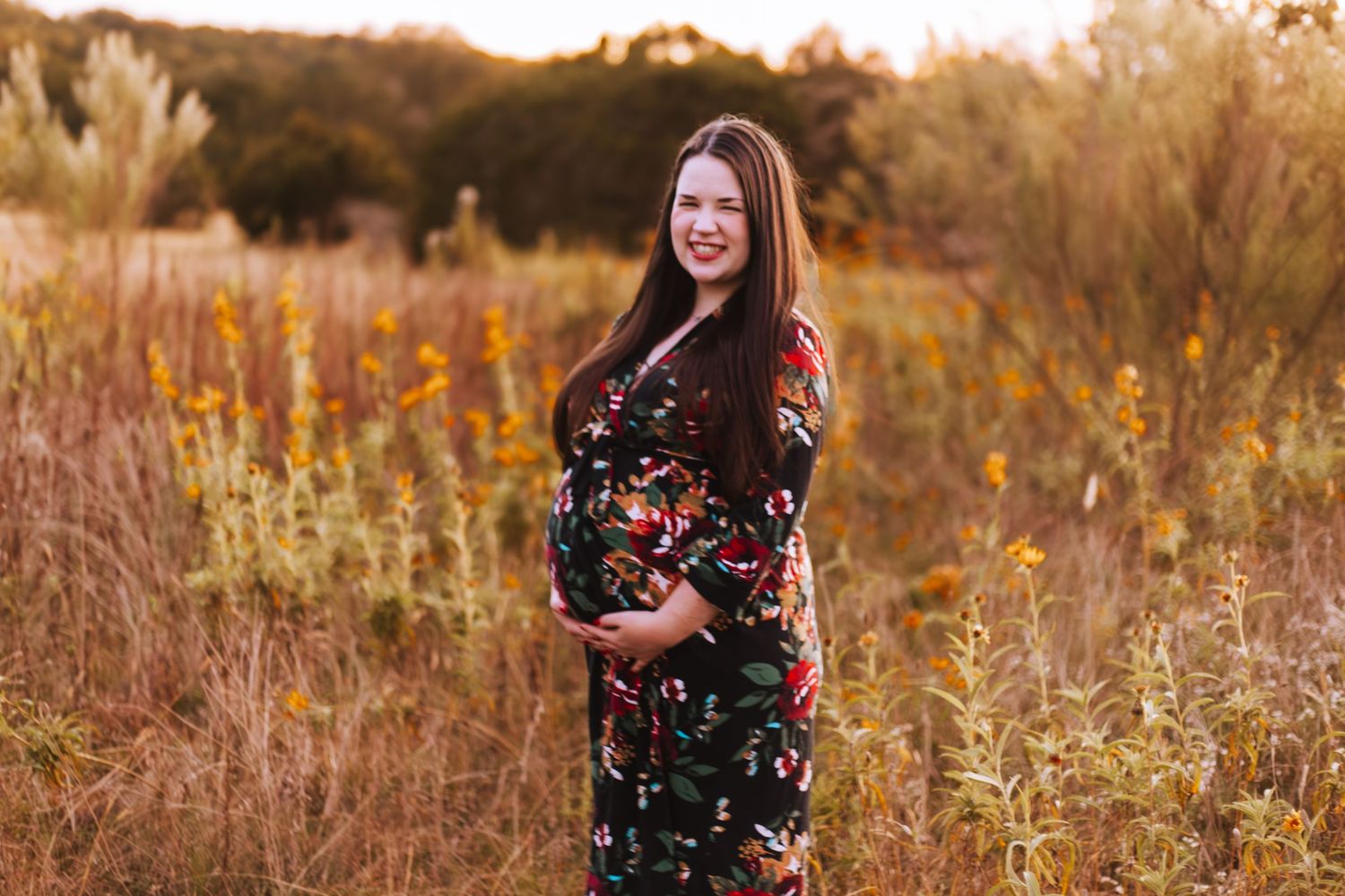A maternity photo in a fall field shows a person wearing a floral dress at sunset with golden grasses in the background.