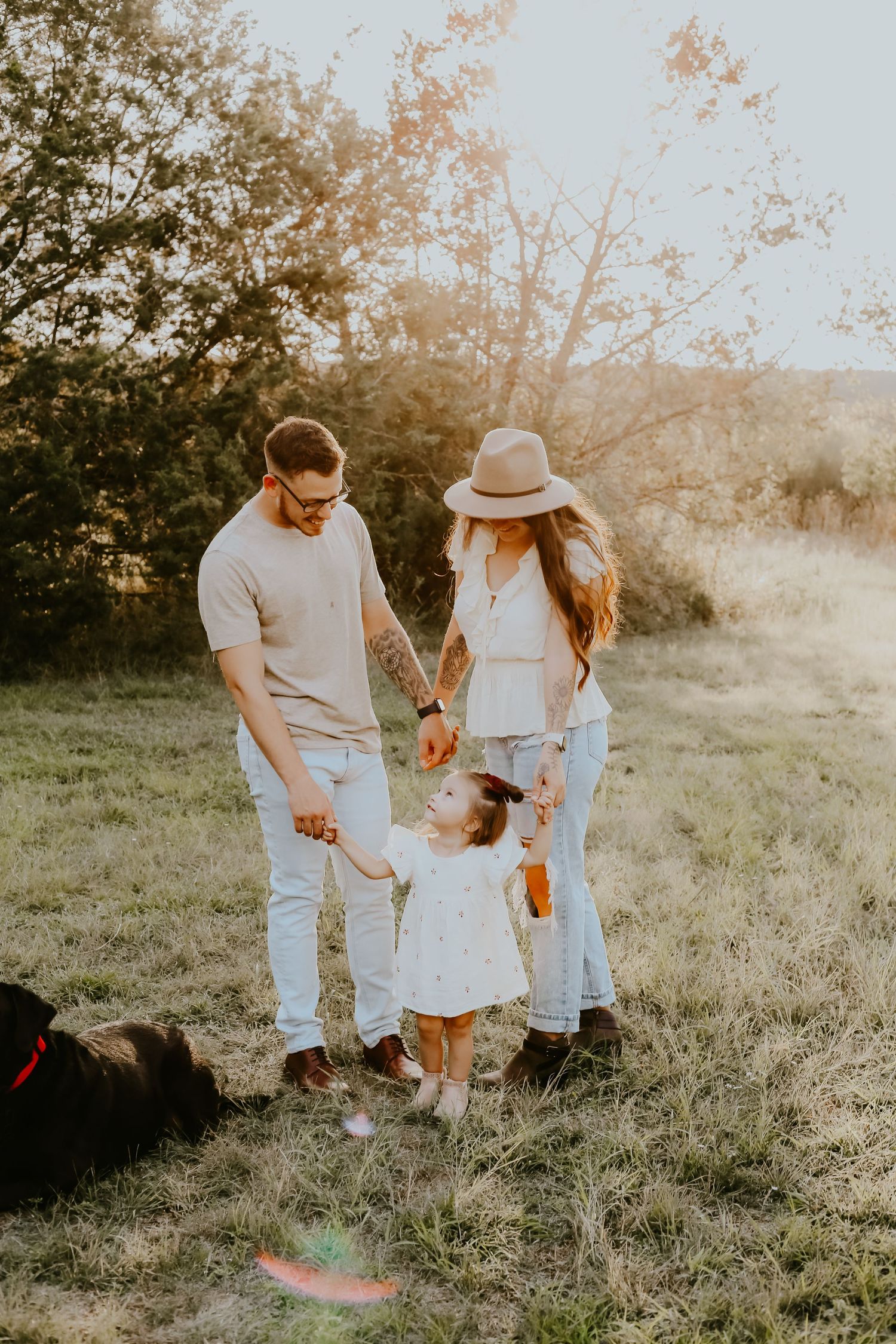 A family holding hands together in a sunlit meadow at sunset wearing coordinated white and neutral outfits.