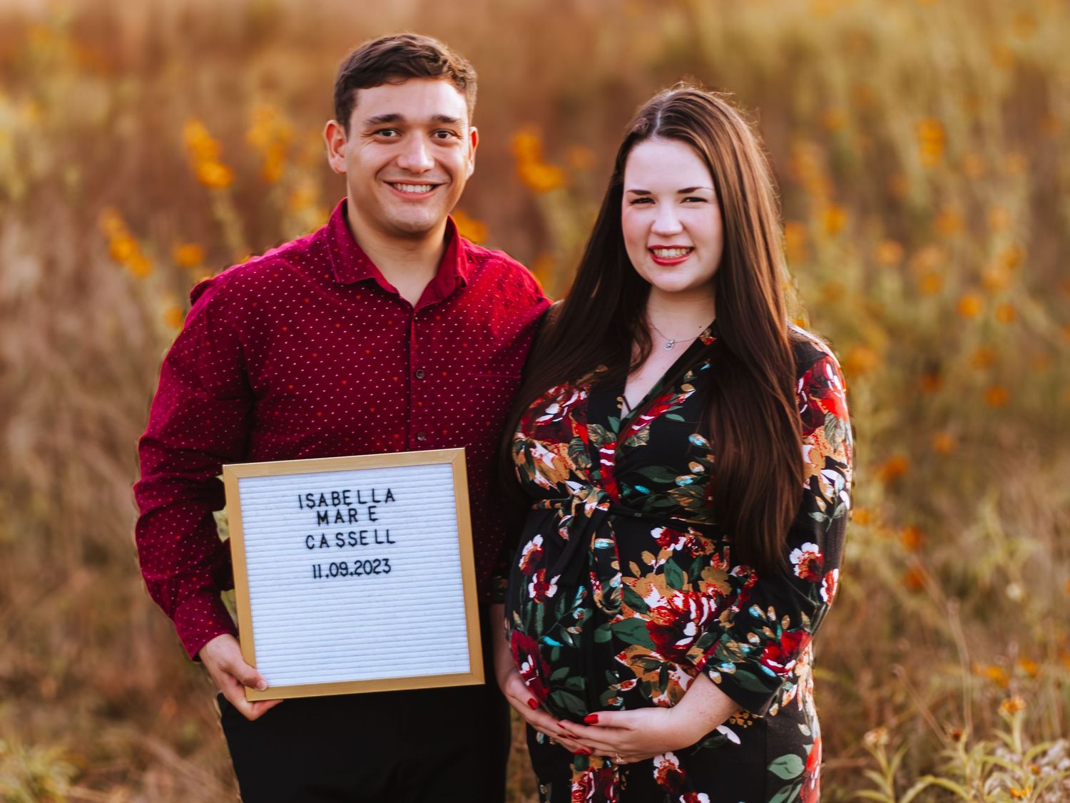 Couple in burgundy and floral attire poses together in autumn field holding announcement signs.