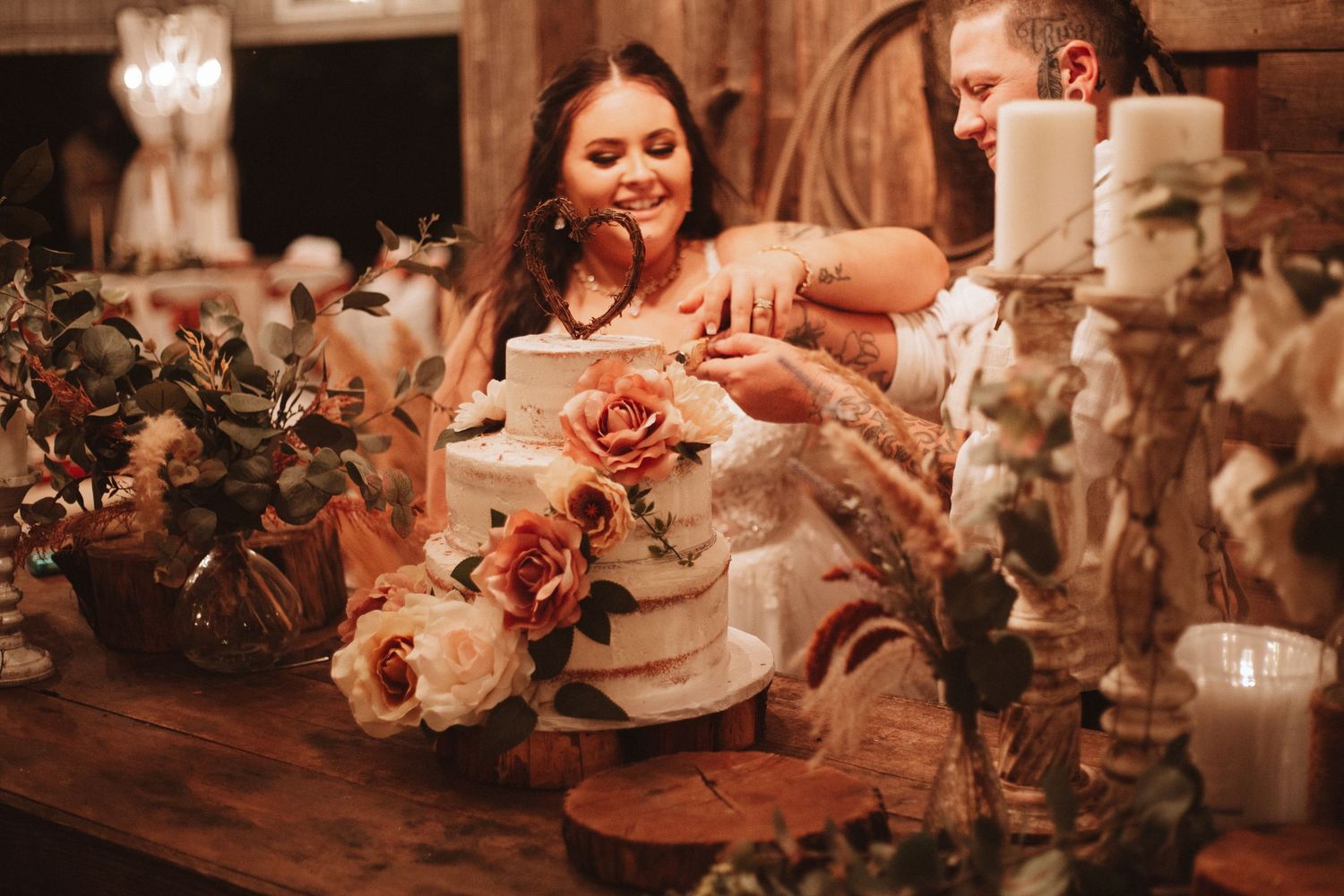 Someone decorates a rustic wedding cake with fresh roses on a wooden table surrounded by candles and floral arrangements.