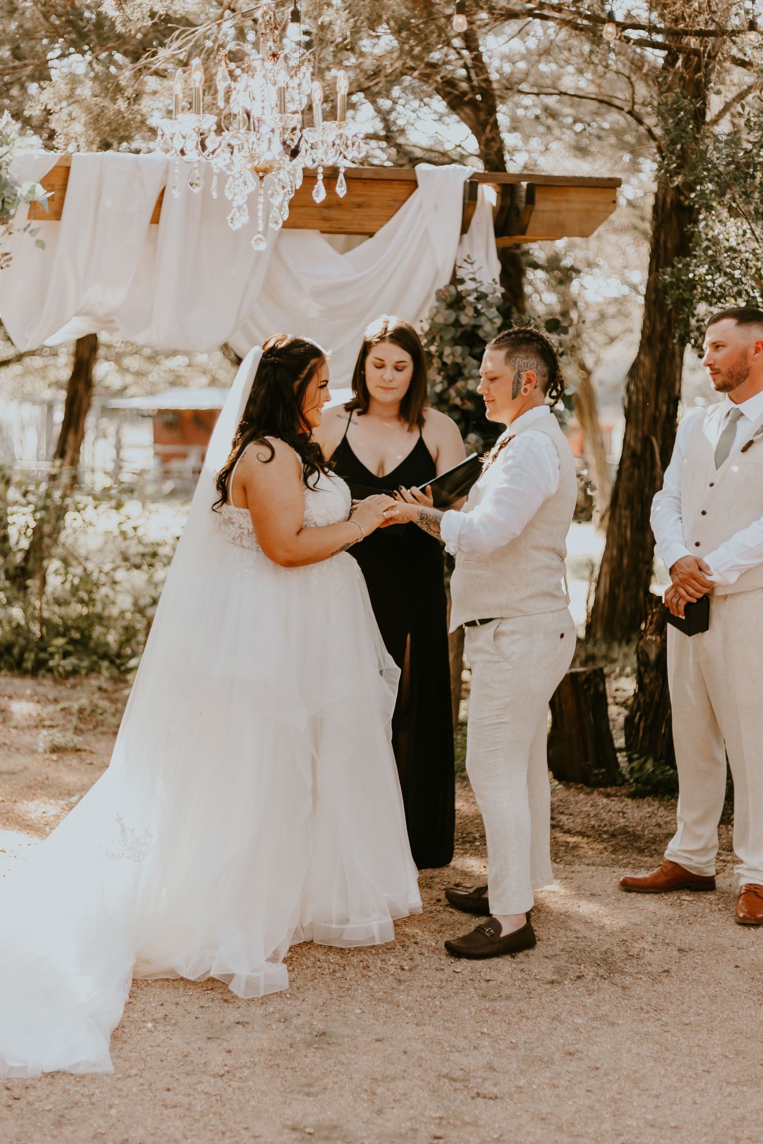 A rustic outdoor wedding ceremony takes place under draped fabric and hanging decorations in a natural setting.