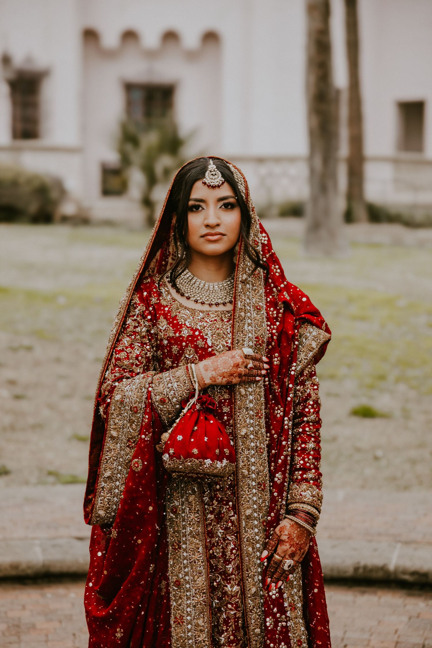 Traditional South Asian bride wearing ornate red and gold embroidered lengha with jewelry and dupatta against castle backdrop.