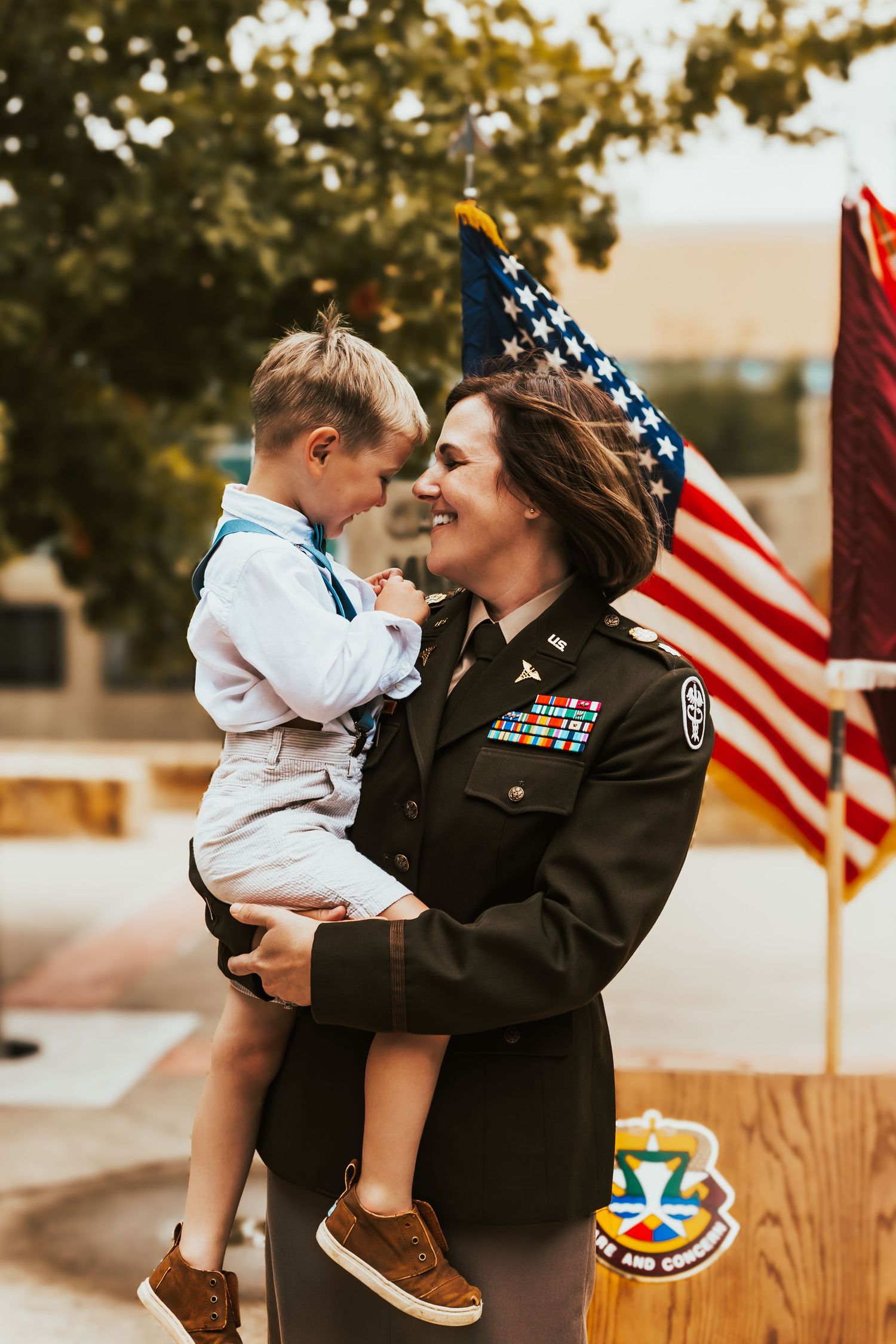 A military service member in dress uniform holds a young child beside an American flag at an outdoor gathering.