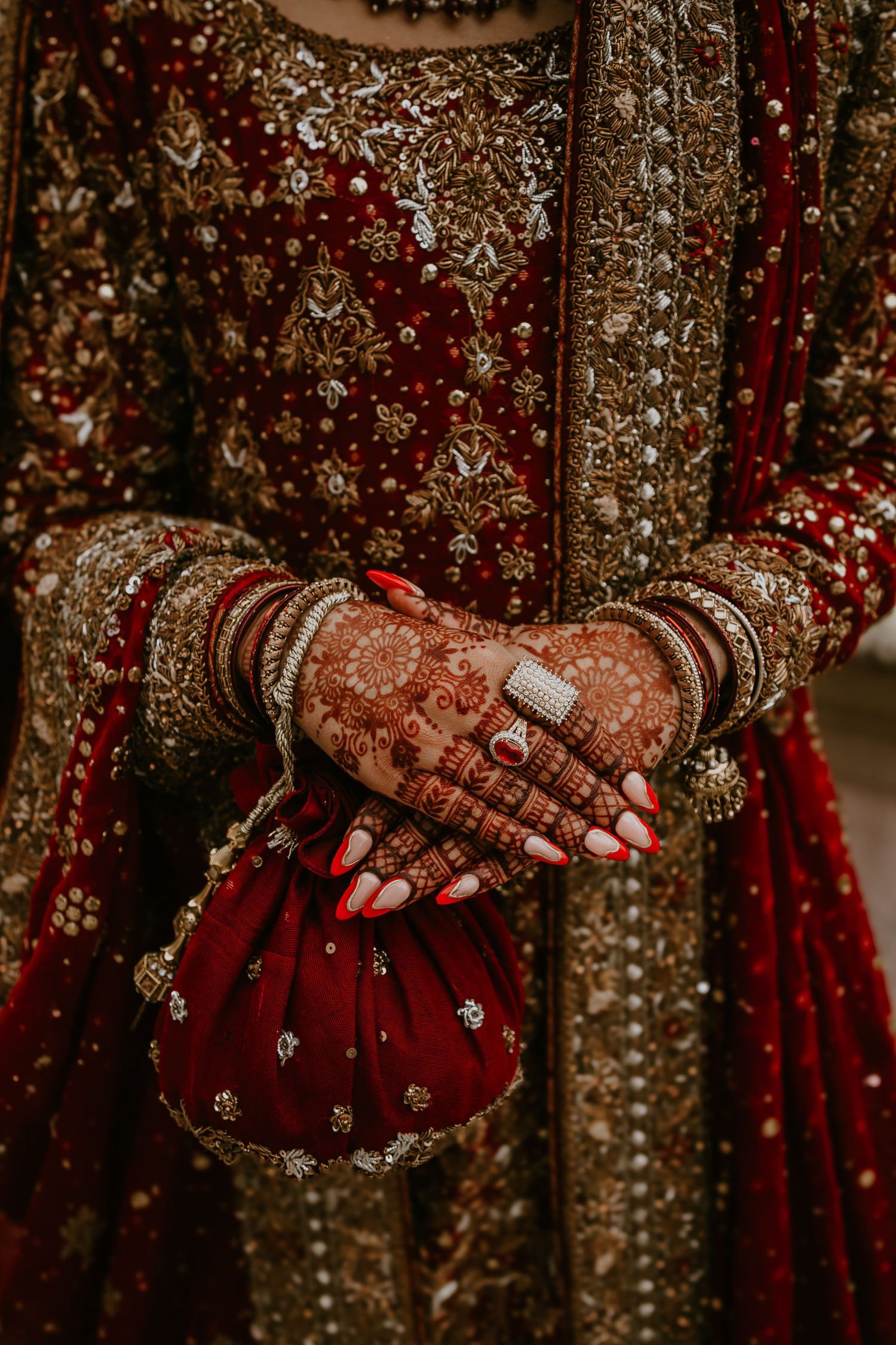 Close-up of henna adorned hands with rings and bangles against an ornate red and gold traditional bridal lehenga dress.