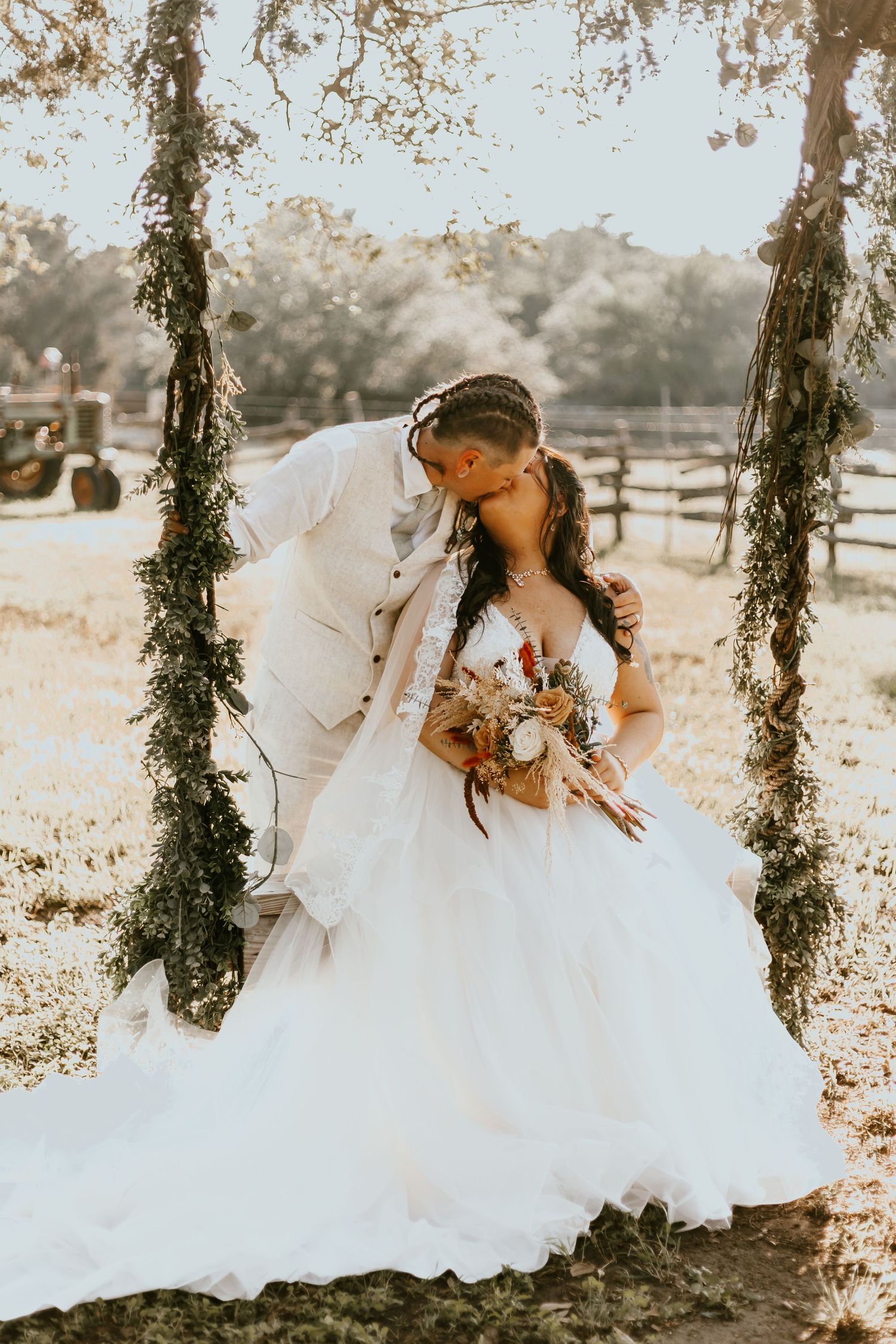A romantic couple shares a moment on a rustic vine-covered swing holding a floral bouquet in dreamy sunlit scenery.