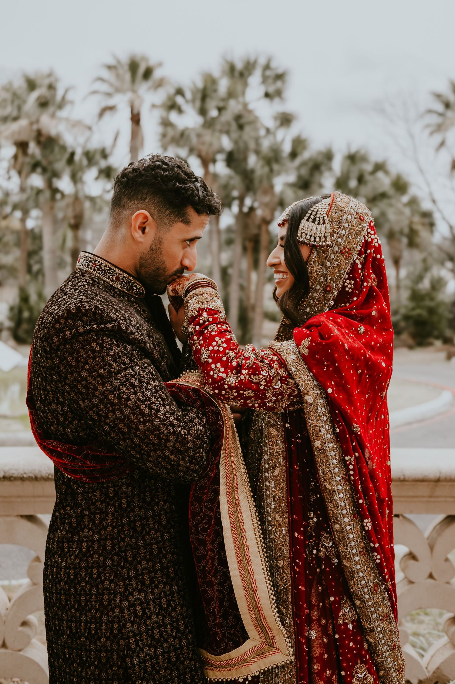 Traditional South Asian wedding couple in ornate burgundy and red attire embrace under palm trees during outdoor ceremony.