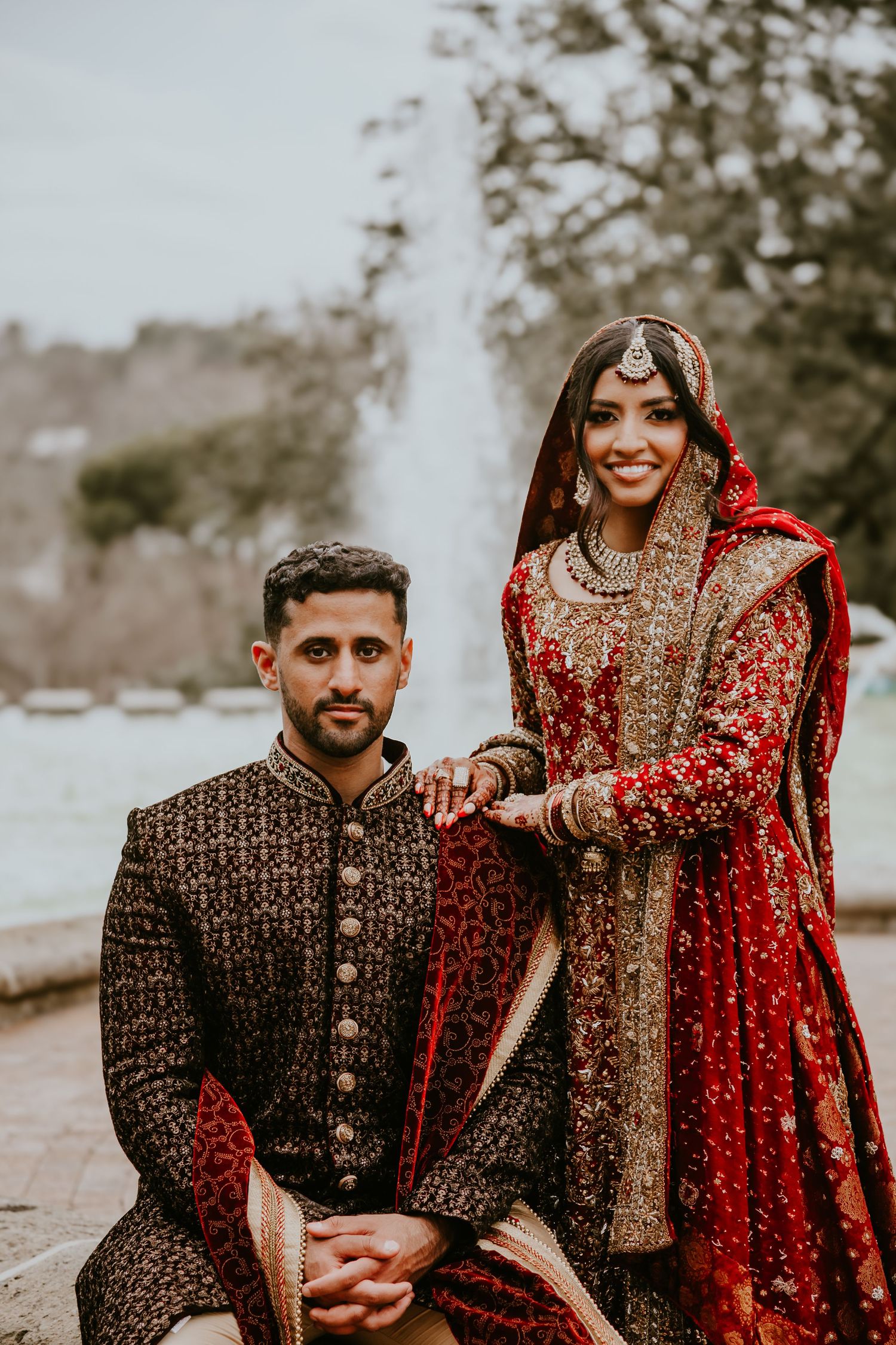 Couple in traditional South Asian wedding attire featuring an ornate red lehenga and dark embroidered sherwani.