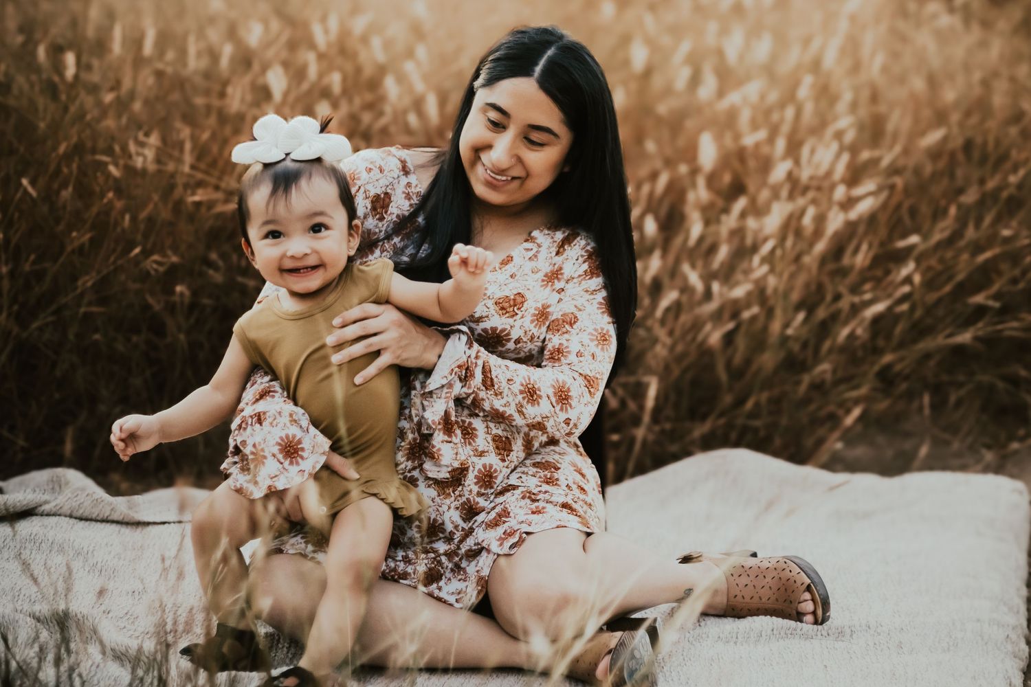 Mother and child in floral dresses sitting together in tall grass during golden hour.