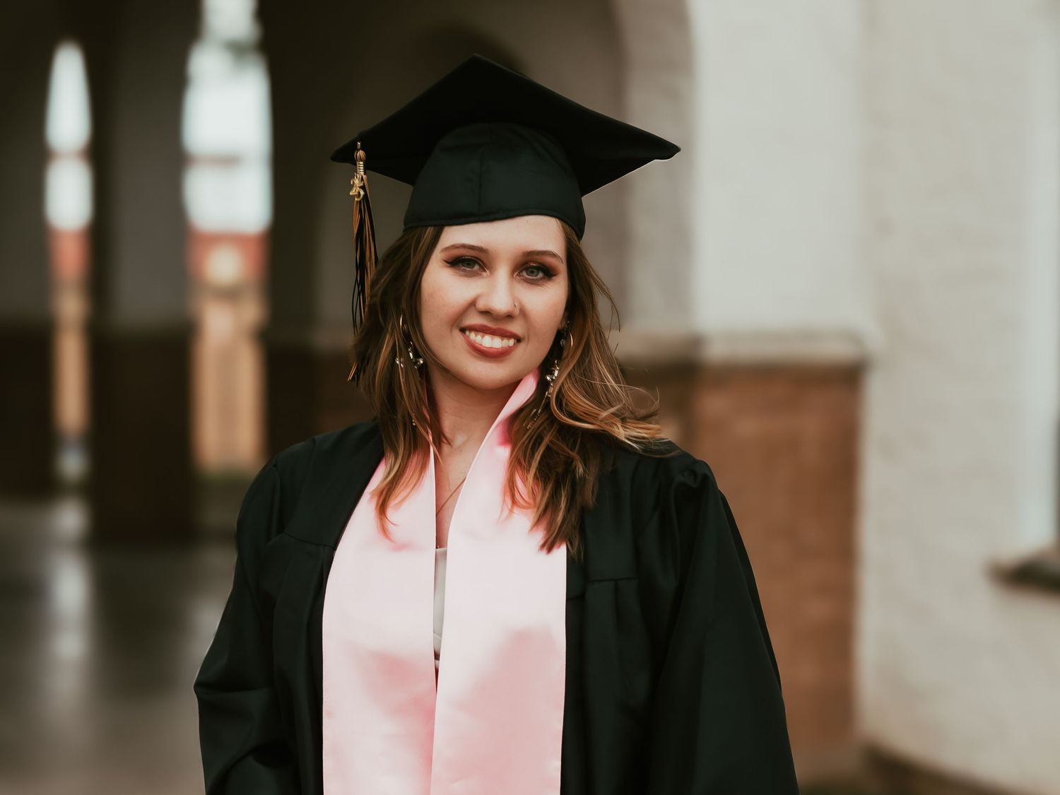 Graduation portrait with black cap and gown against warm-toned architectural background.