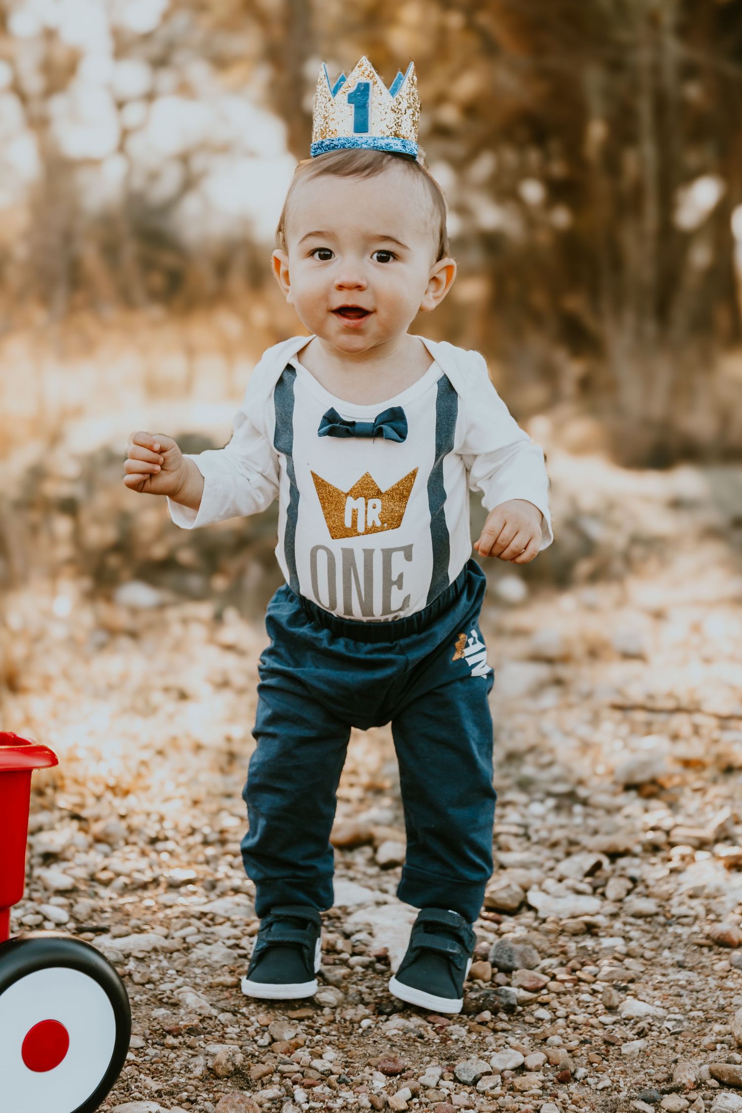 Baby wearing blue crown, suspenders and bow tie outfit stands near red wagon on autumn day.