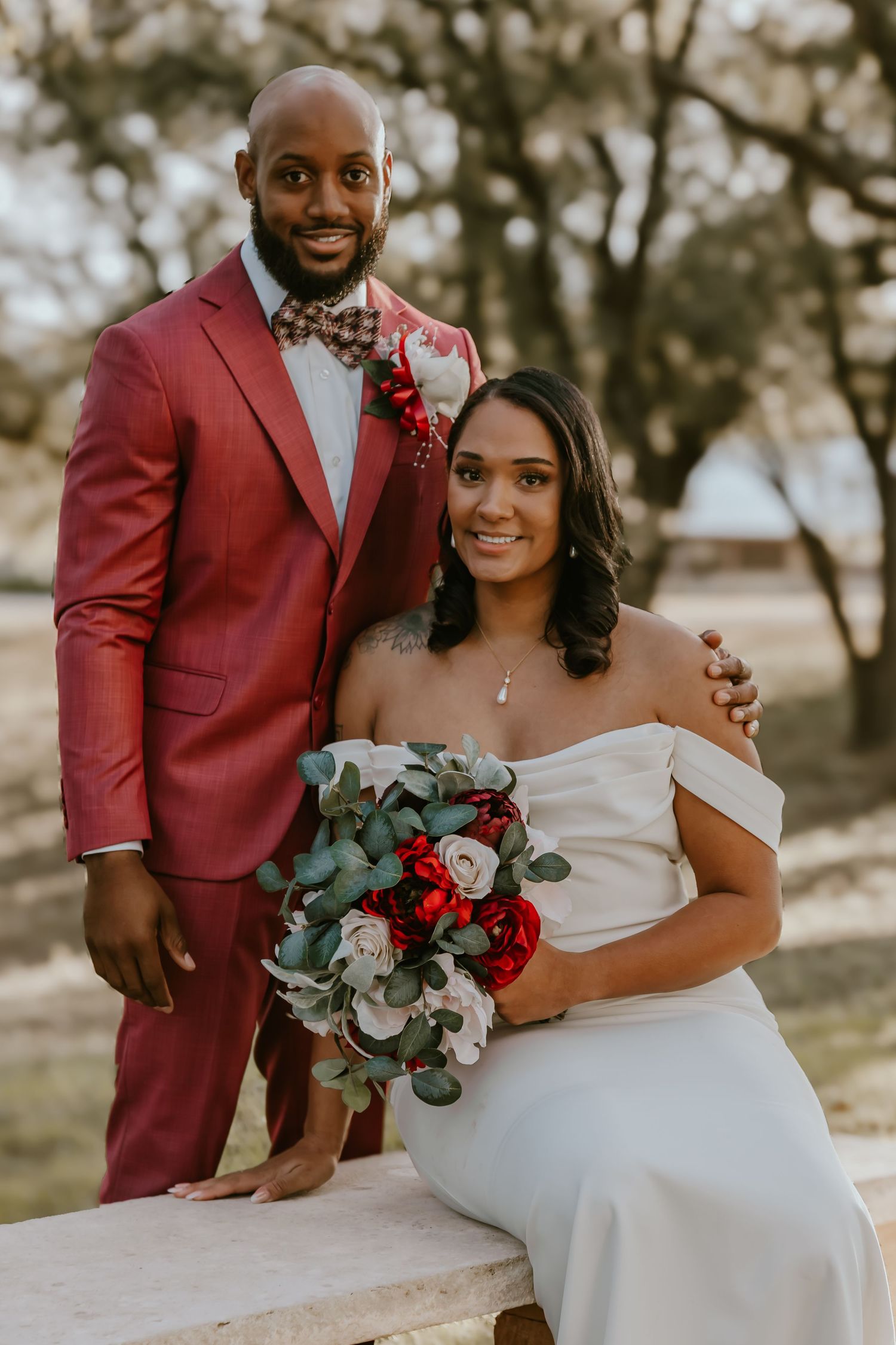A couple in wedding attire sits and embraces outdoors, with the groom in burgundy suit and bride in white gown.