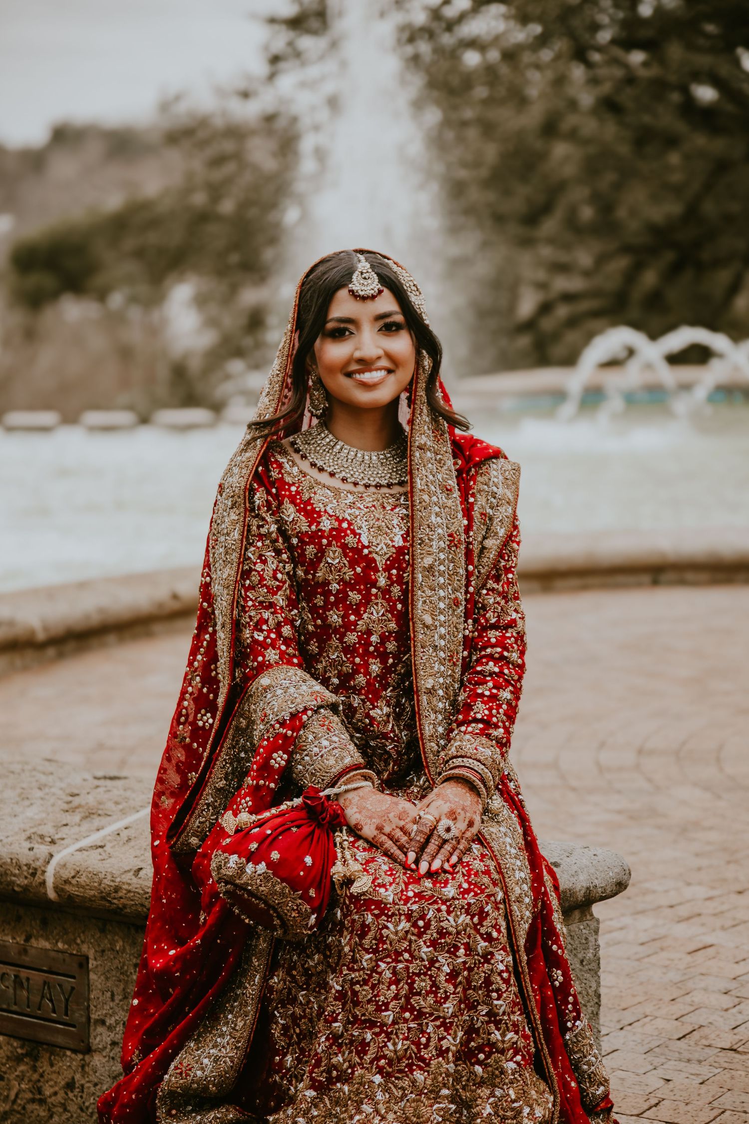 South Asian bride wearing ornate red and gold traditional bridal lehenga sits on stone wall in outdoor garden setting.