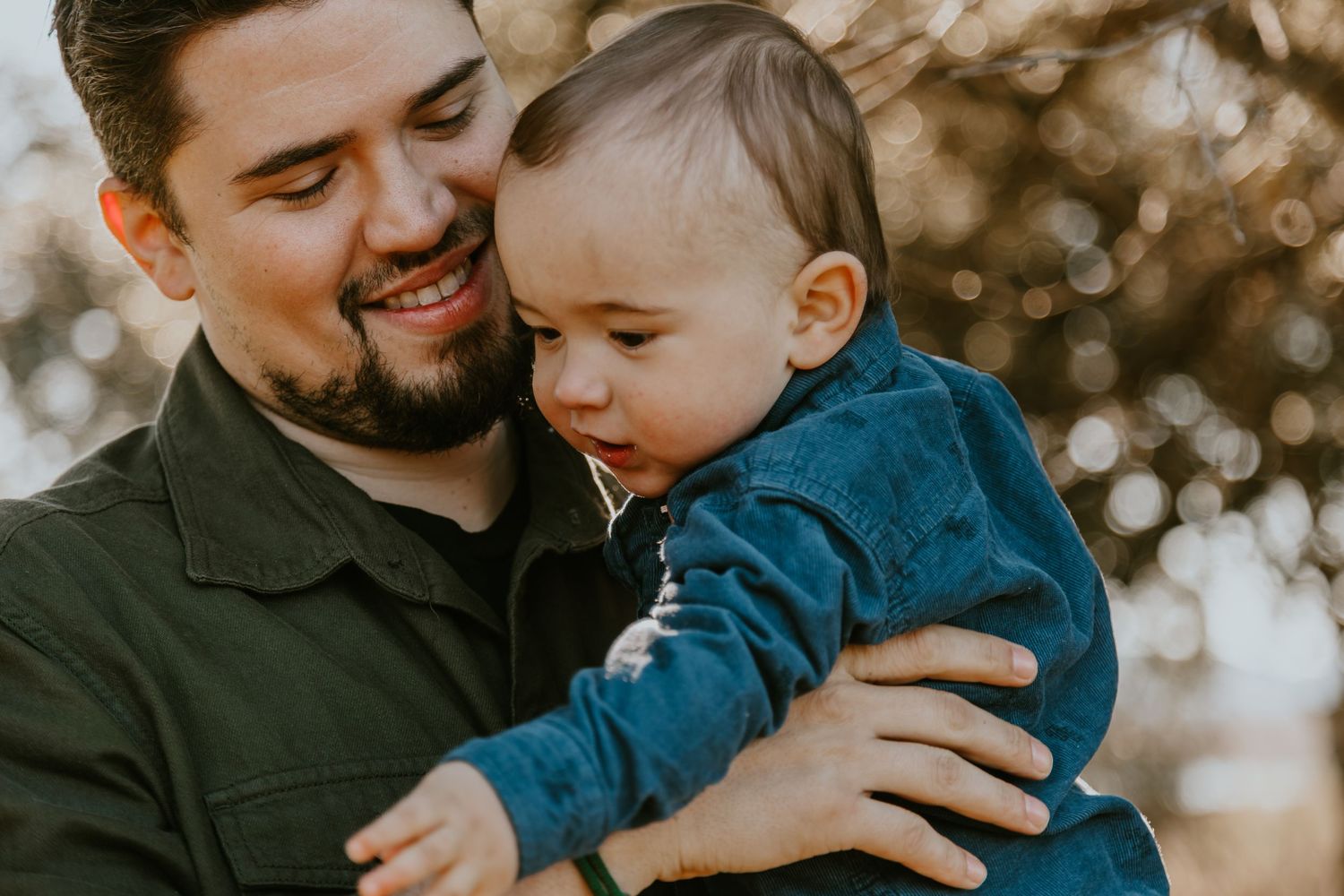 A bearded man in a black jacket holds and smiles warmly at a baby wearing a blue sweater in front of bokeh lights.