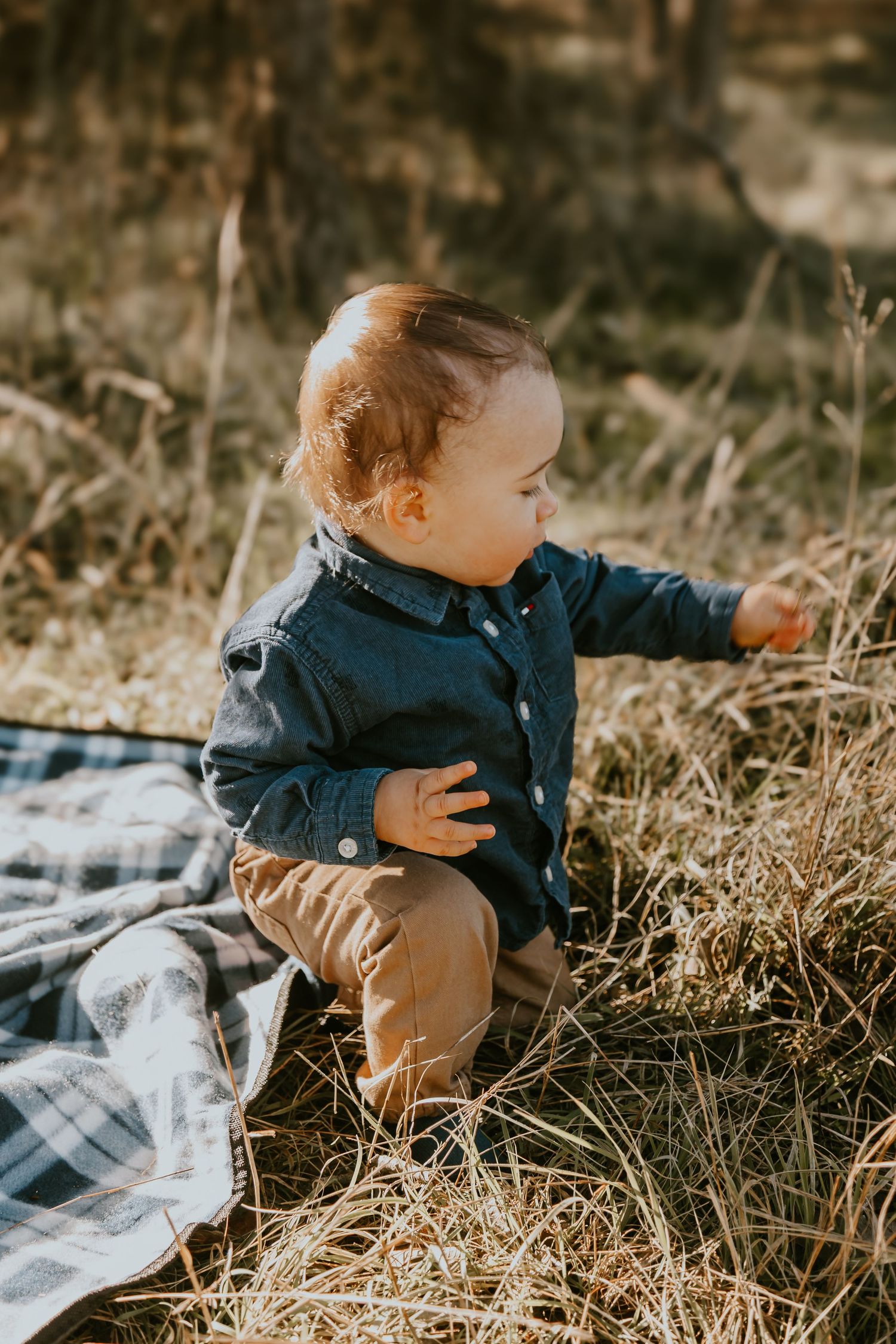 Small toddler in navy button-up shirt and khaki pants sits on plaid blanket in golden autumn field in warm sunlight.
