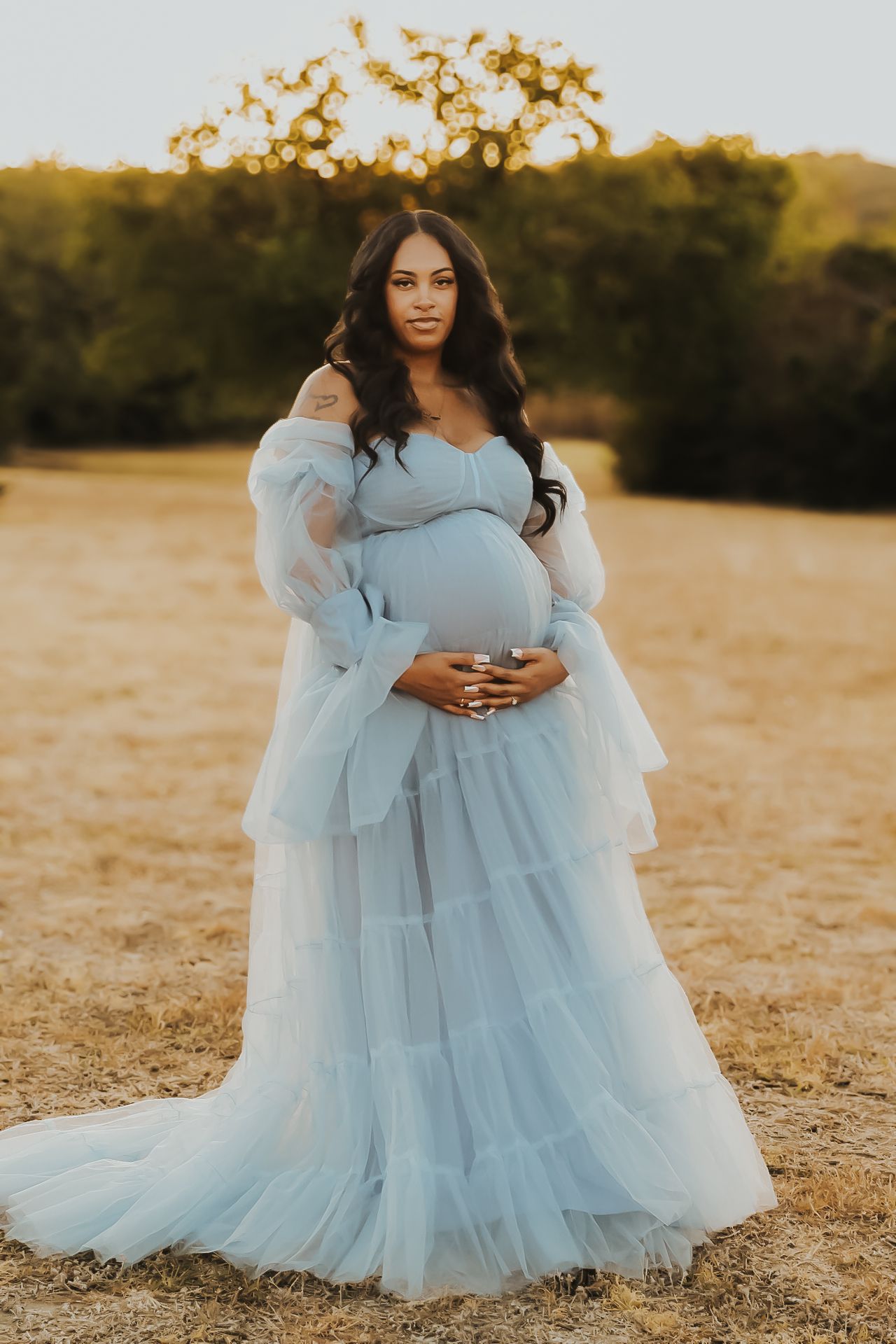 Ethereal maternity portraits in a flowing light blue tulle gown against a golden sunset in an open field.