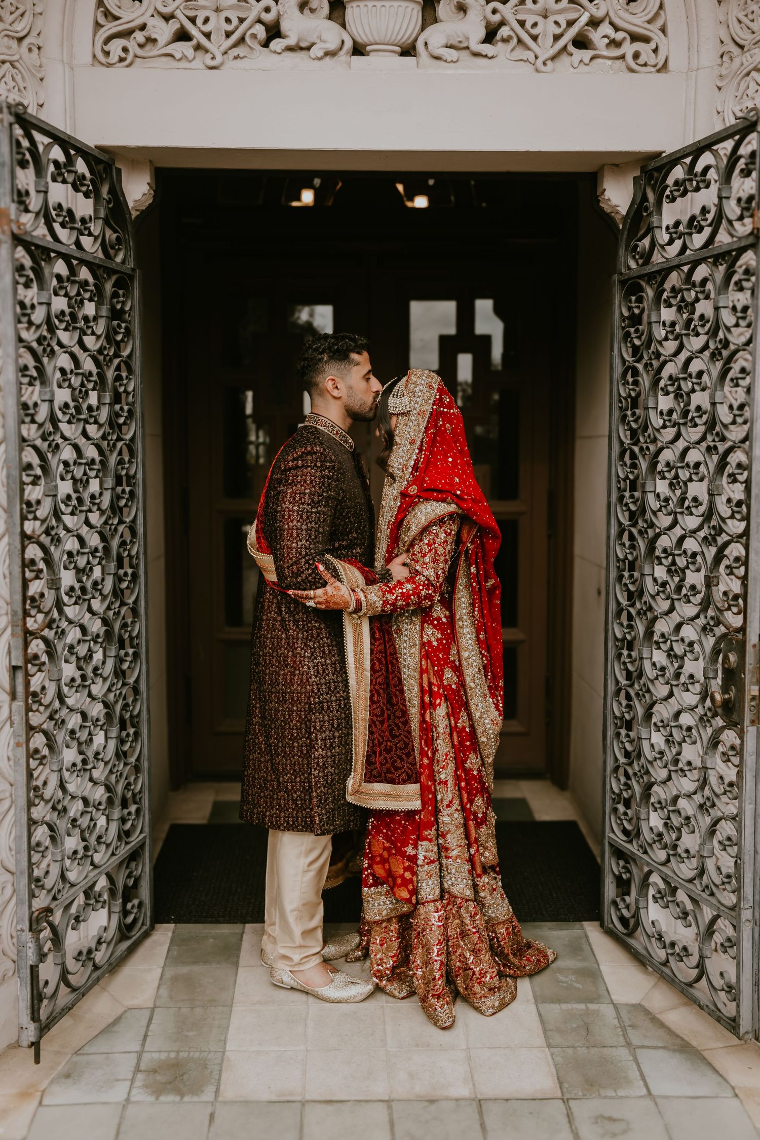 Traditional Indian wedding couple in ornate red and brown outfits stand in an intricately carved doorway.