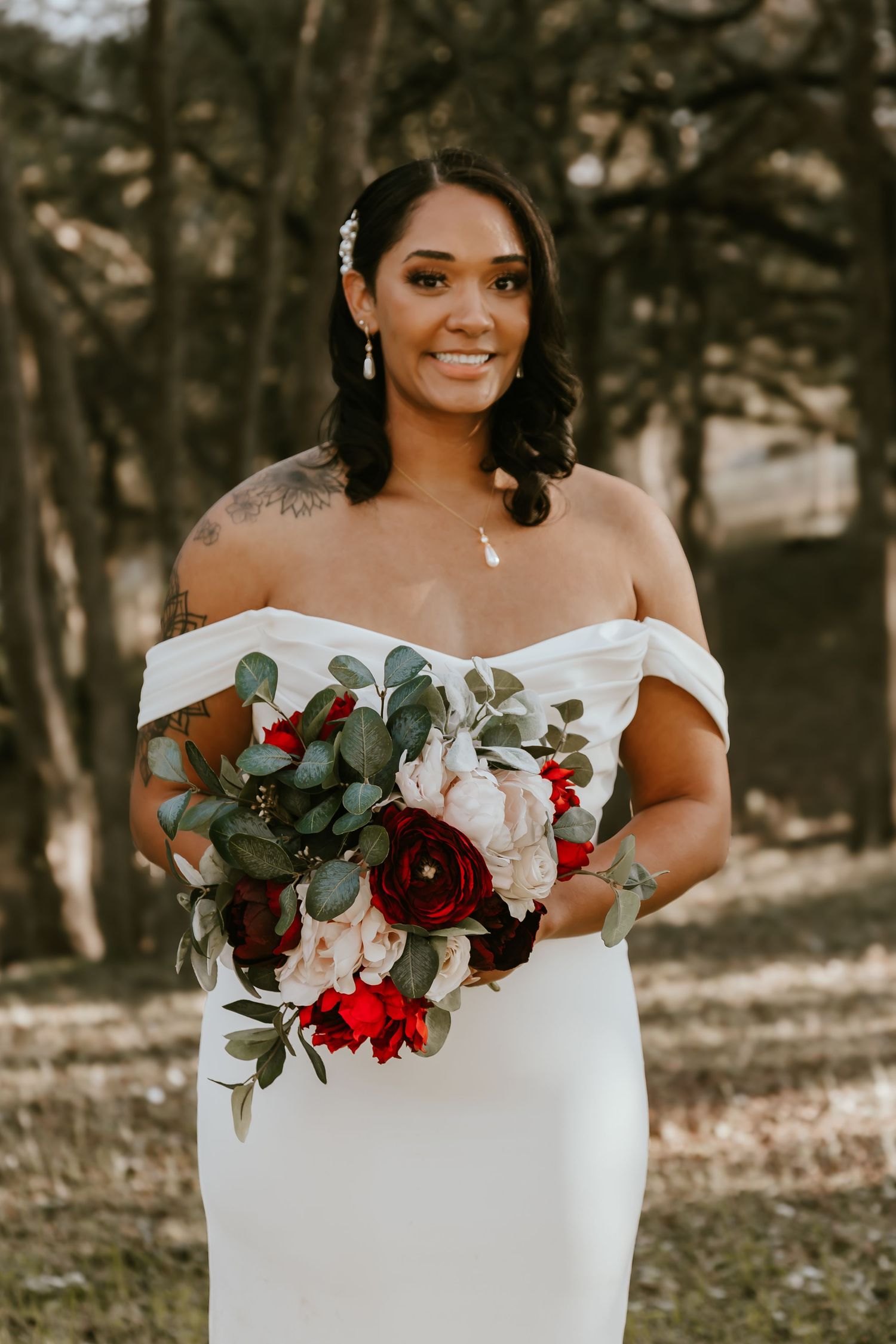 A bride in an off-shoulder white wedding gown holds a beautiful bouquet with burgundy and white flowers and eucalyptus.