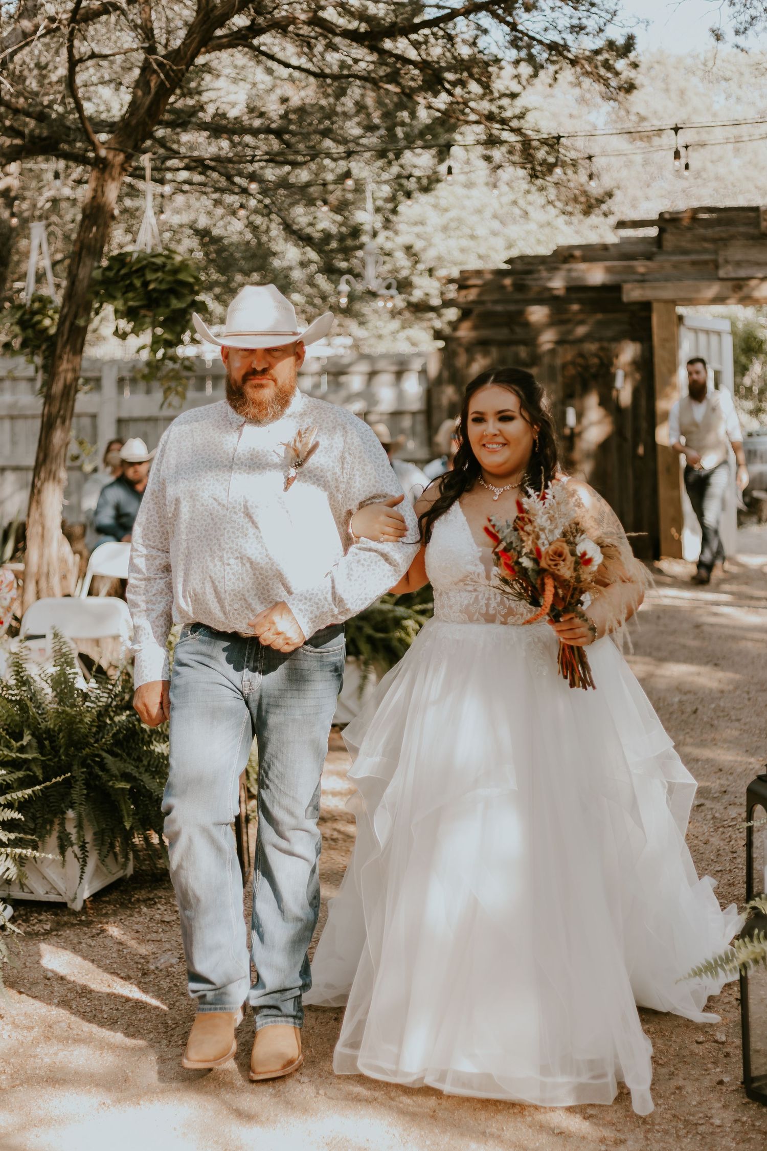 A bride in a flowing white gown walks down a sunlit outdoor path with a companion in a white cowboy hat and jeans.