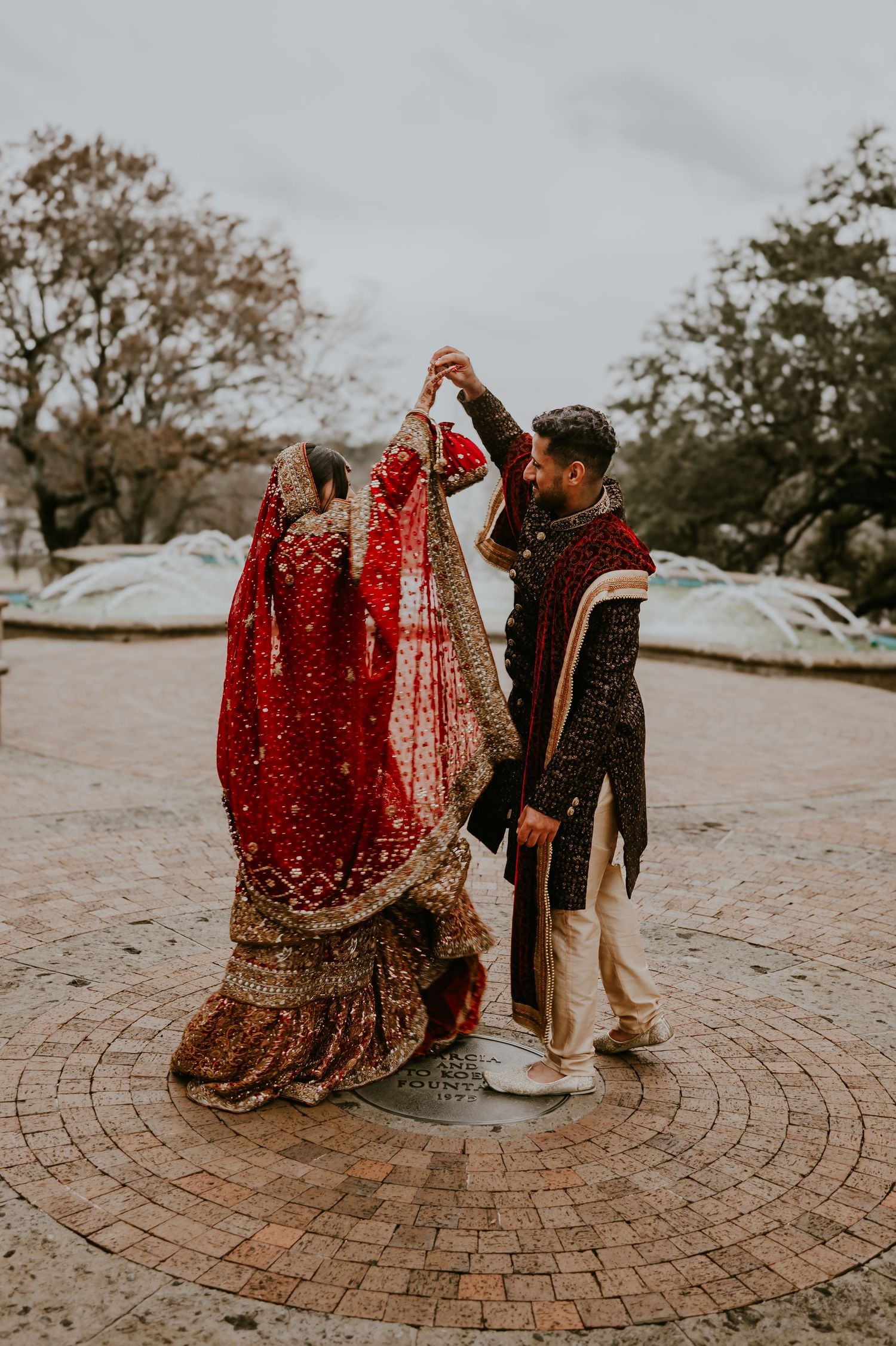 A couple in traditional Indian wedding attire dances together on a circular brick patio during an outdoor celebration.
