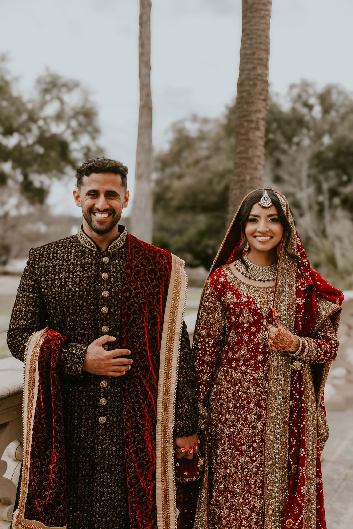 A couple dressed in ornate traditional South Asian wedding attire featuring deep red and black embroidered outfits standing outdoors.
