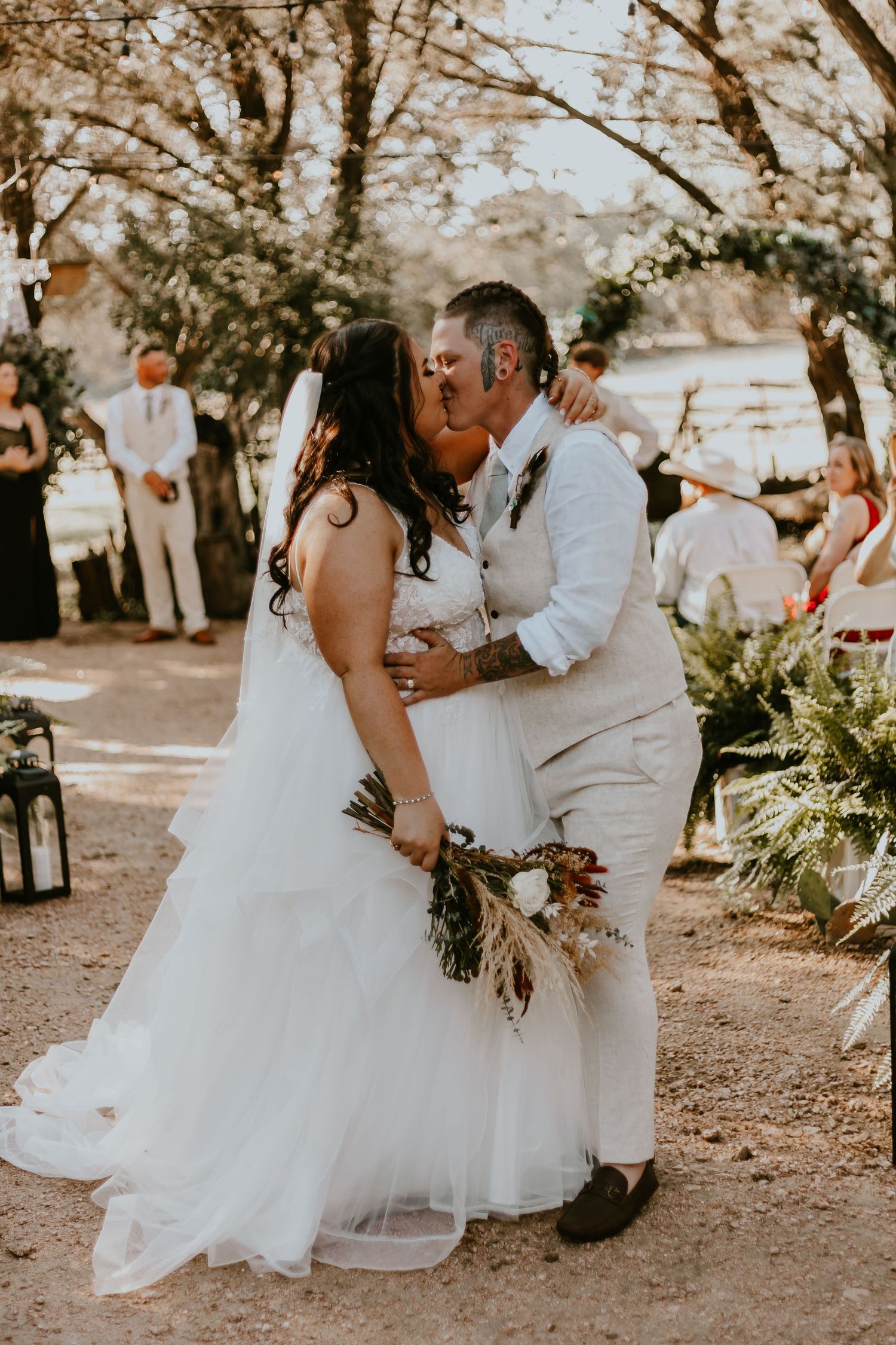 A romantic outdoor wedding scene with a couple sharing their first kiss during a rustic ceremony under trees at sunset.