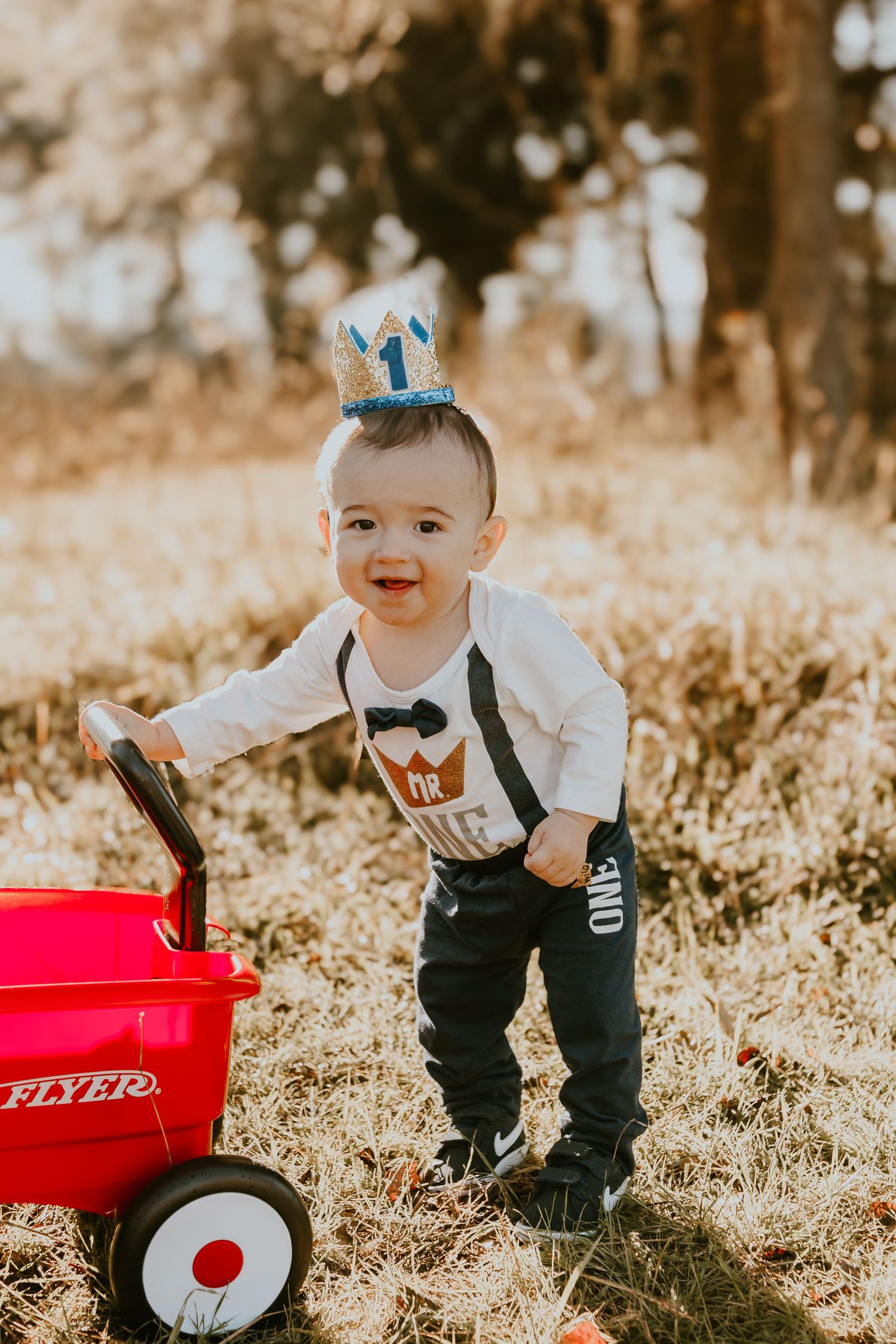 Happy baby wearing birthday crown and suspenders smiles while playing with red Radio Flyer wagon in sunny outdoor setting.