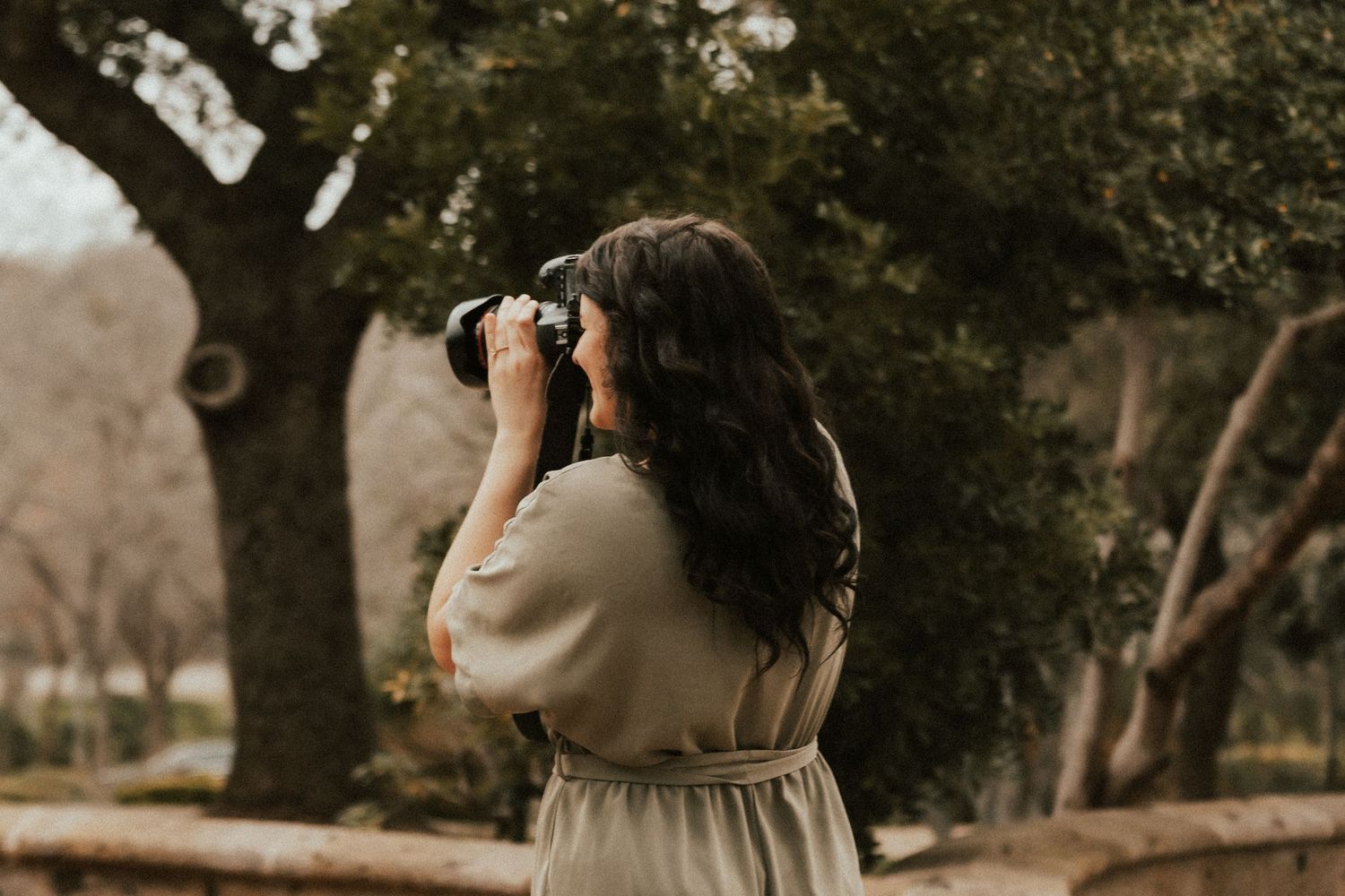 Photographer in cream dress captures photos in a natural setting with trees.