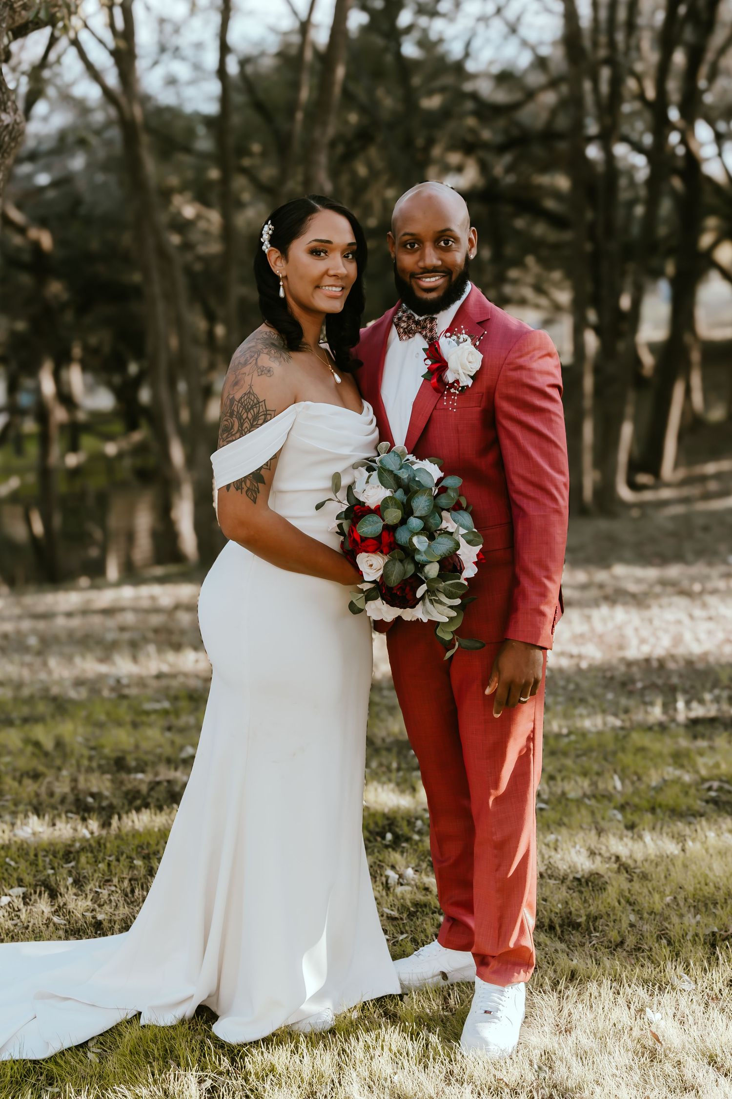 Wedding couple in elegant white dress and coral suit pose with bouquet in outdoor setting.