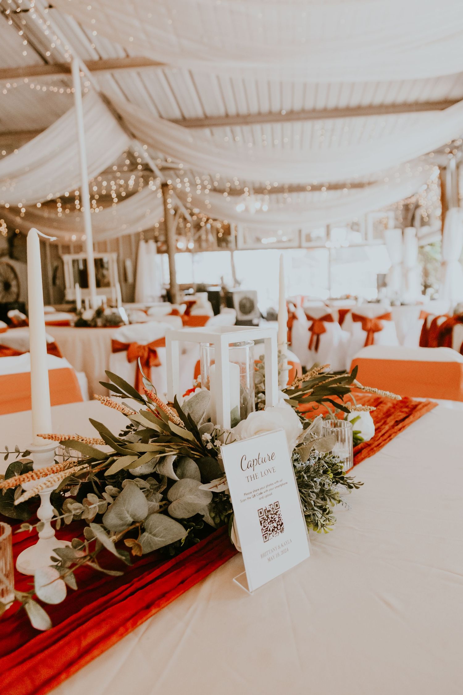 Wedding reception table decorated with white lanterns, red accents, and greenery under draped tent ceiling with twinkle lights.