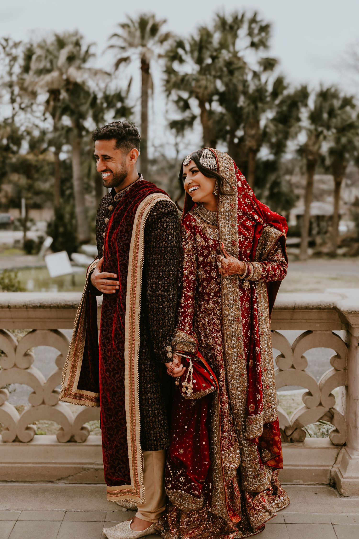 Traditional South Asian wedding couple in ornate red and gold attire pose together outdoors among palm trees.