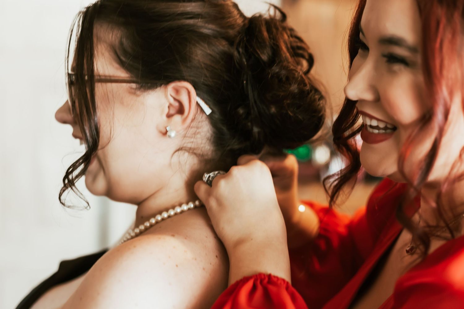 Two friends share a joyful moment together with one wearing a pearl necklace while another has red hair.