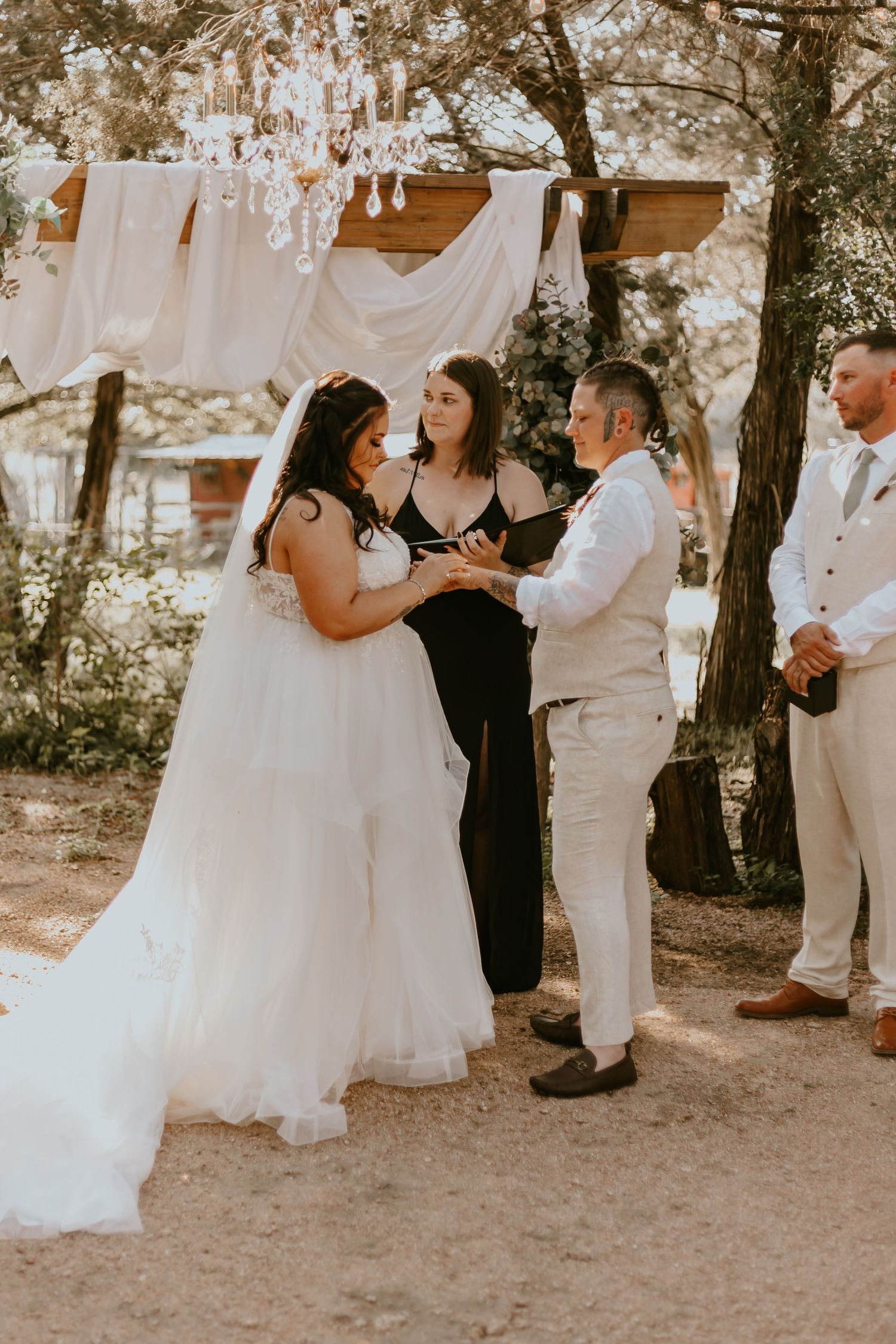 An outdoor wedding ceremony shows attendees gathering beneath trees in a rustic setting with warm sepia tones.