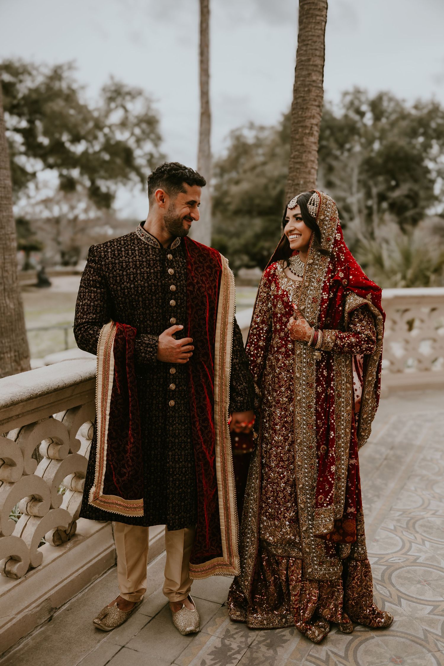 A couple wearing traditional South Asian wedding attire standing together on a terrace with palm trees in the background.