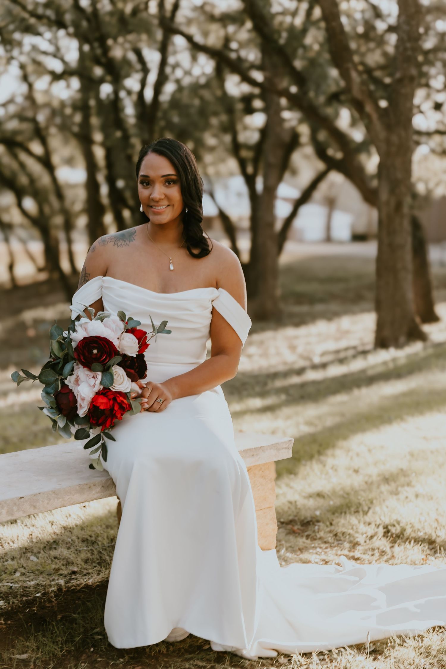 Beautiful outdoor bridal portrait featuring an off-shoulder white gown and burgundy floral bouquet in a sunlit garden setting.