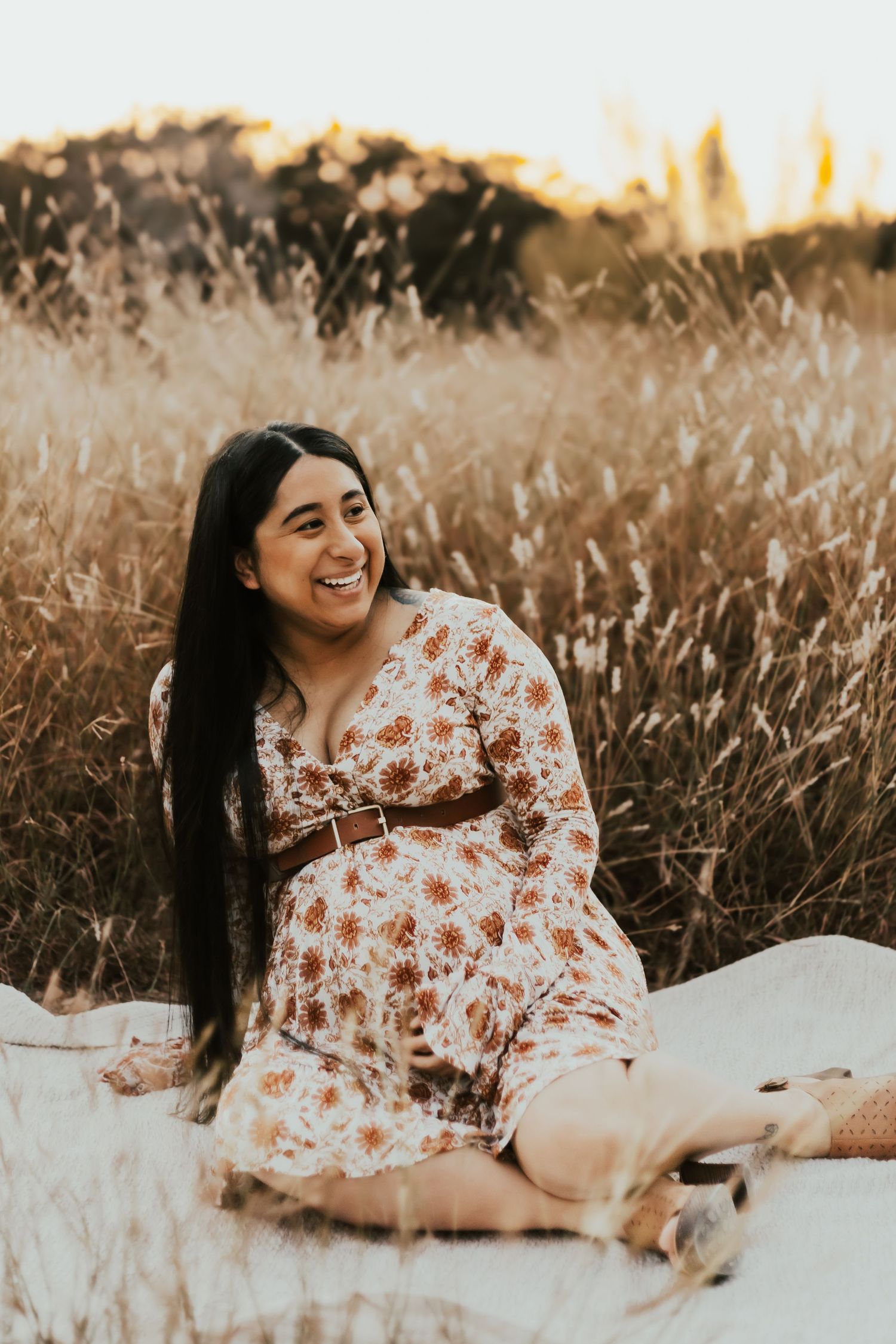 Expecting mother in floral dress laughs joyfully while sitting on white blanket in golden sunlit field at sunset.