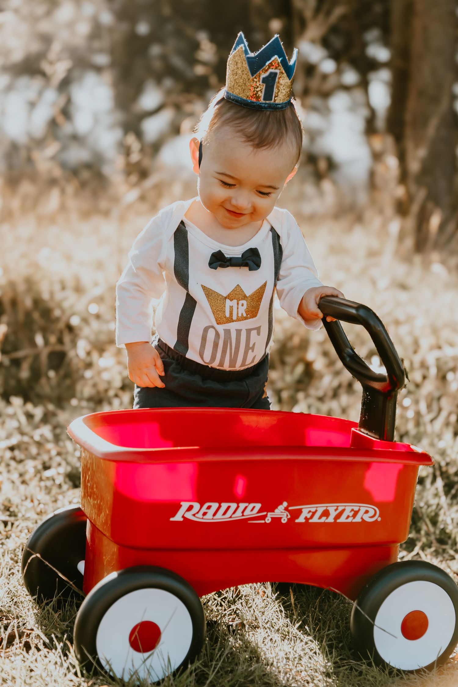 Smiling child wearing crown and suspenders outfit stands next to classic red Radio Flyer wagon in sunlit field.