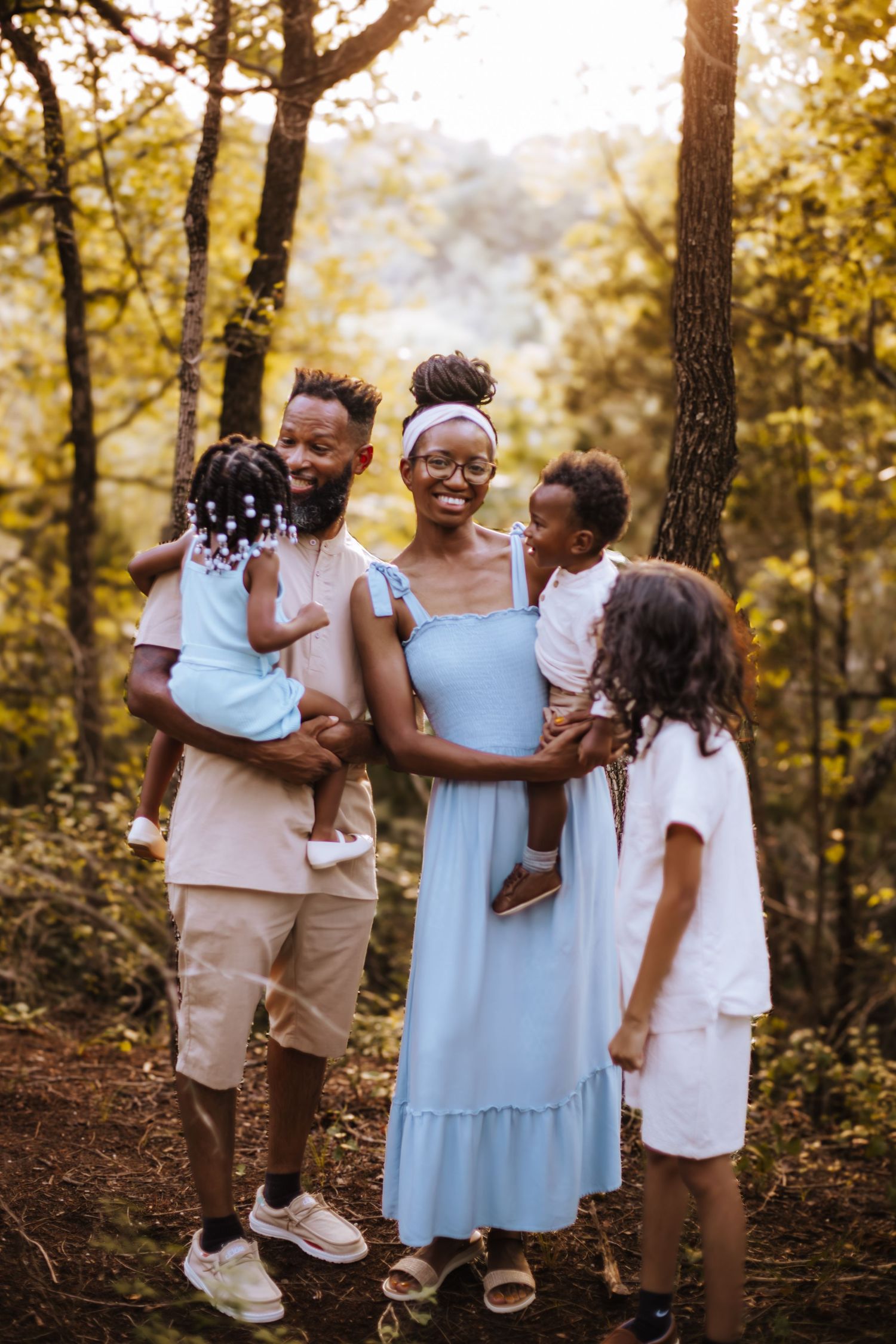 A loving family dressed in soft pastels enjoys tender moments together in a sun-dappled forest setting.