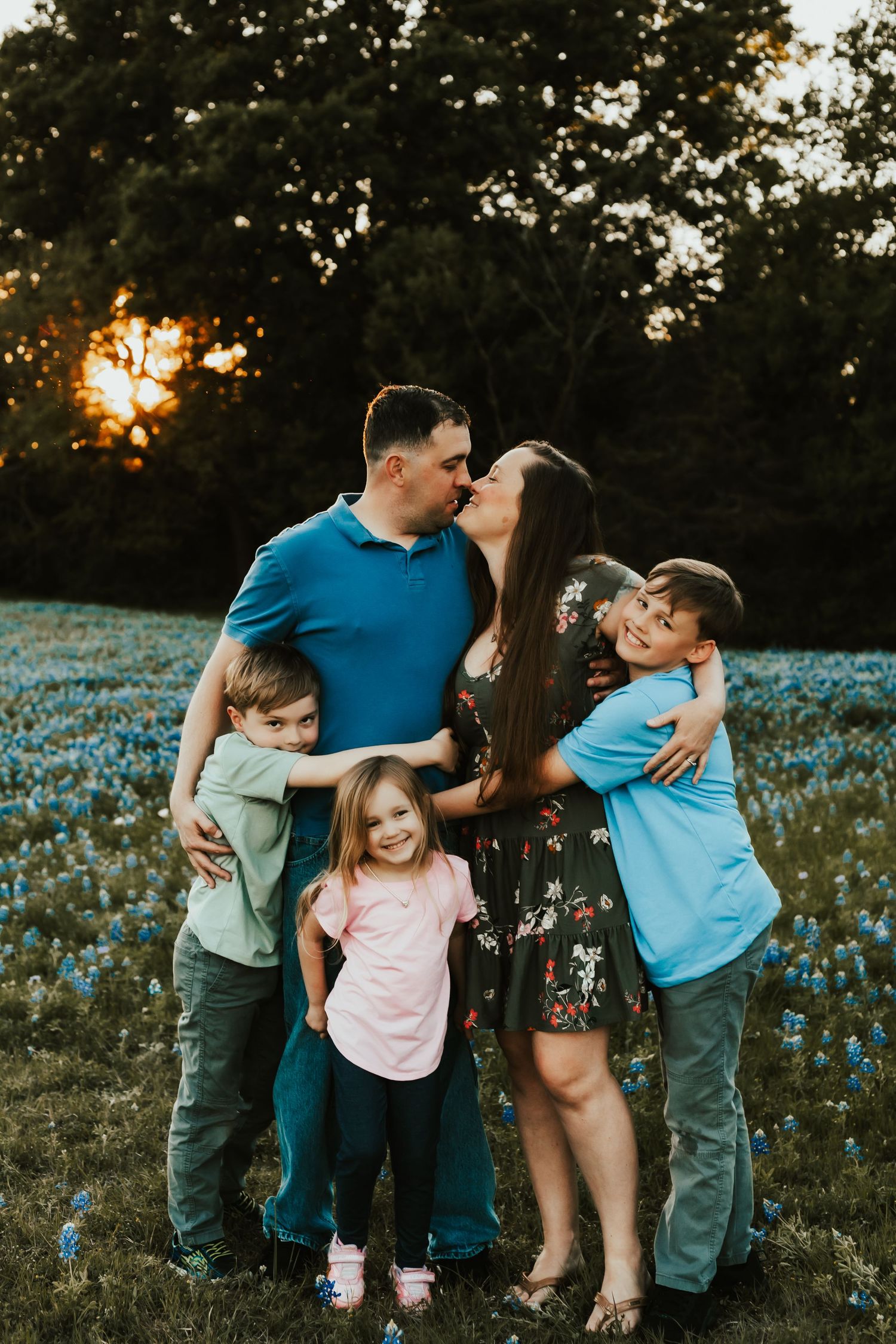 Family stands together in a field of blue wildflowers during golden sunset hour.