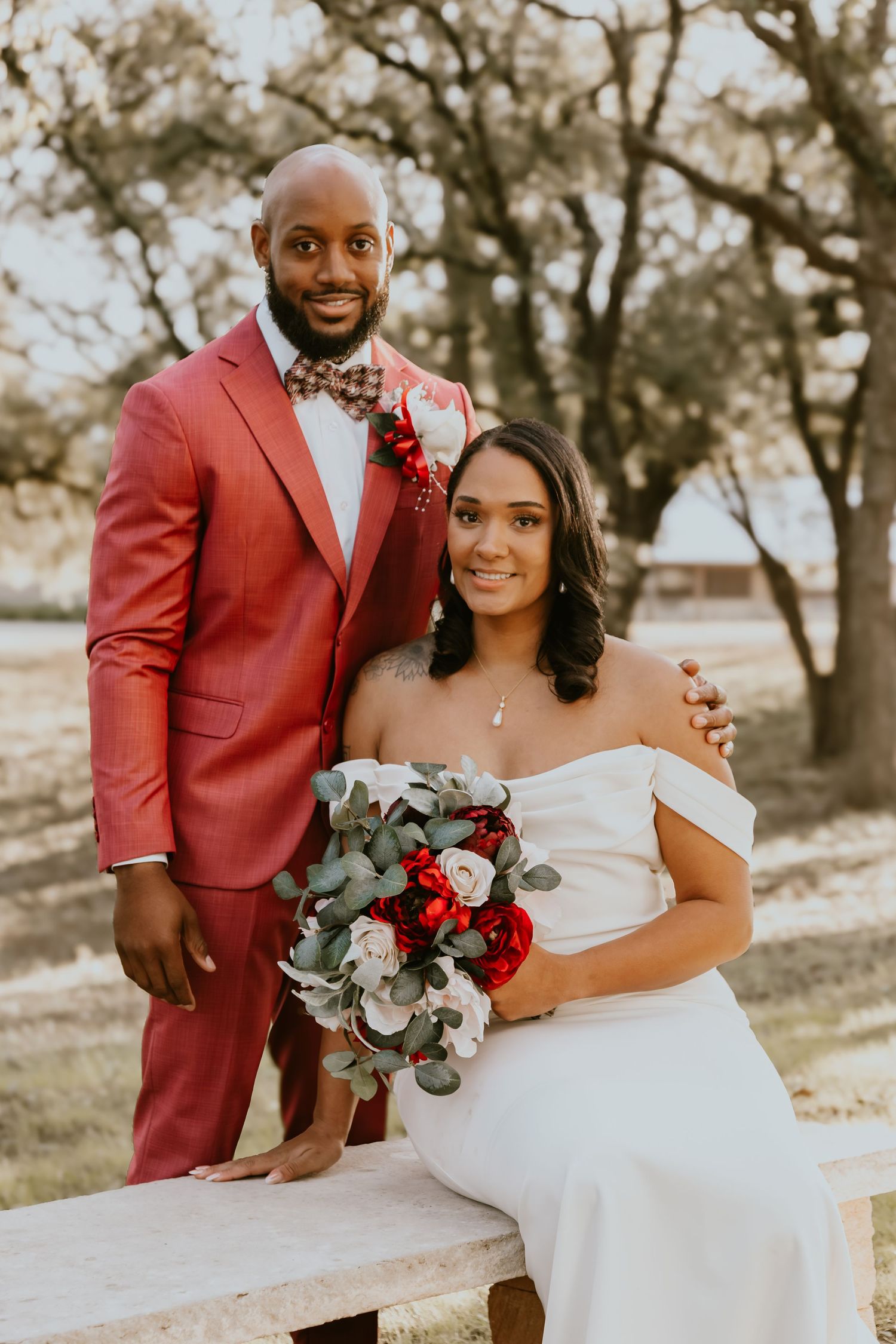 Portrait of a couple in formal wedding attire outside, with red roses and eucalyptus bouquet, against a nature backdrop.