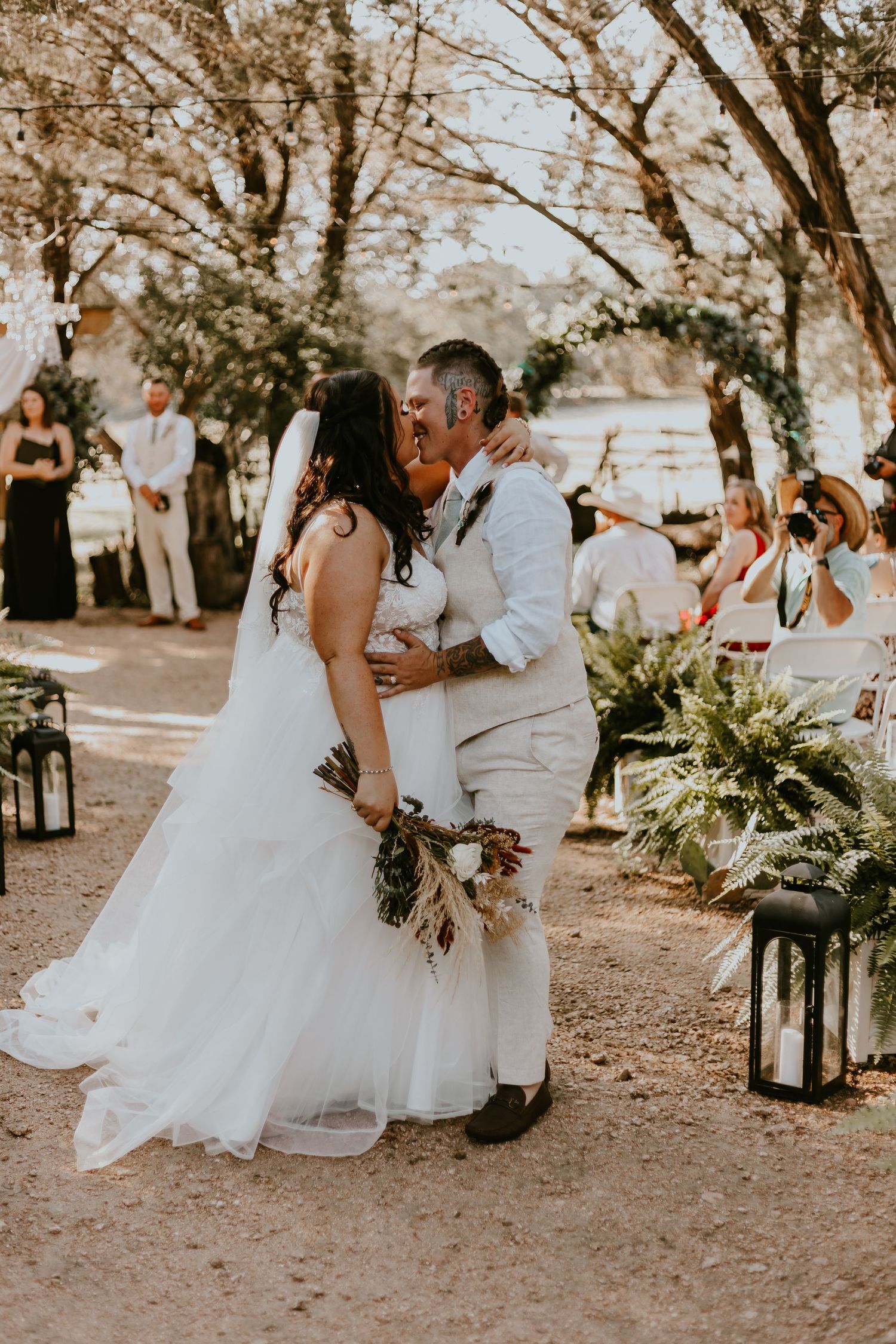 A romantic outdoor wedding moment as newlyweds share a kiss on a sun-dappled path lined with lanterns and ferns.
