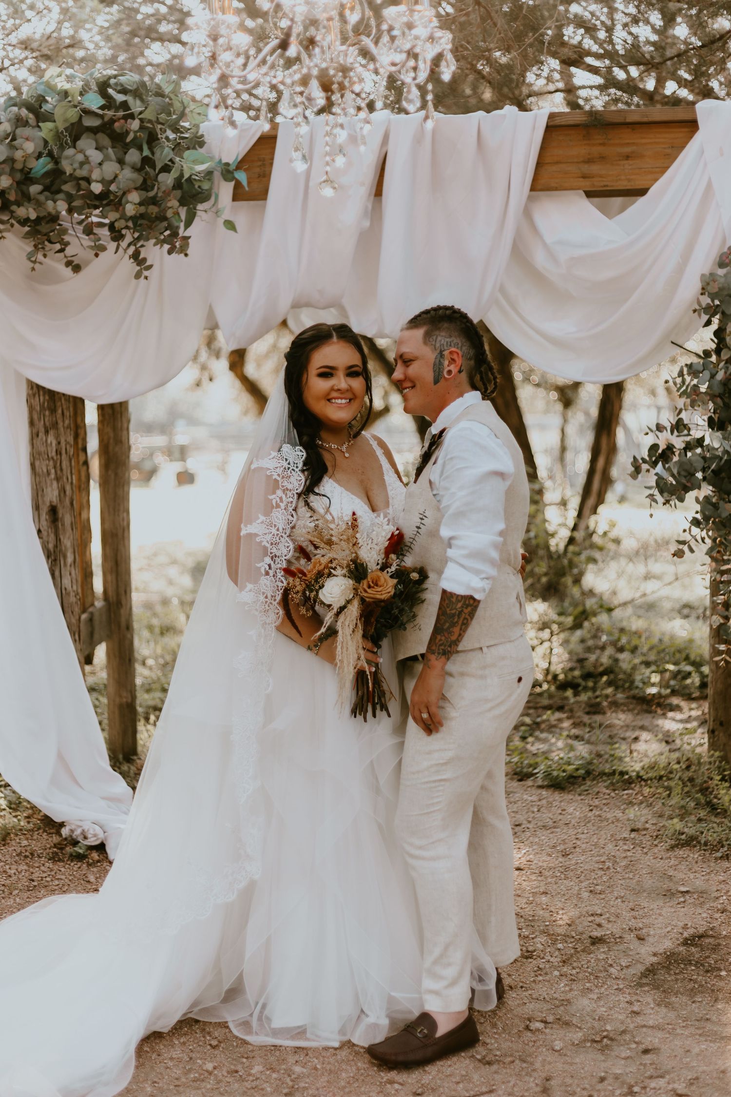 A happy couple poses together on their rustic outdoor wedding day amongst flowy white fabric and natural greenery.