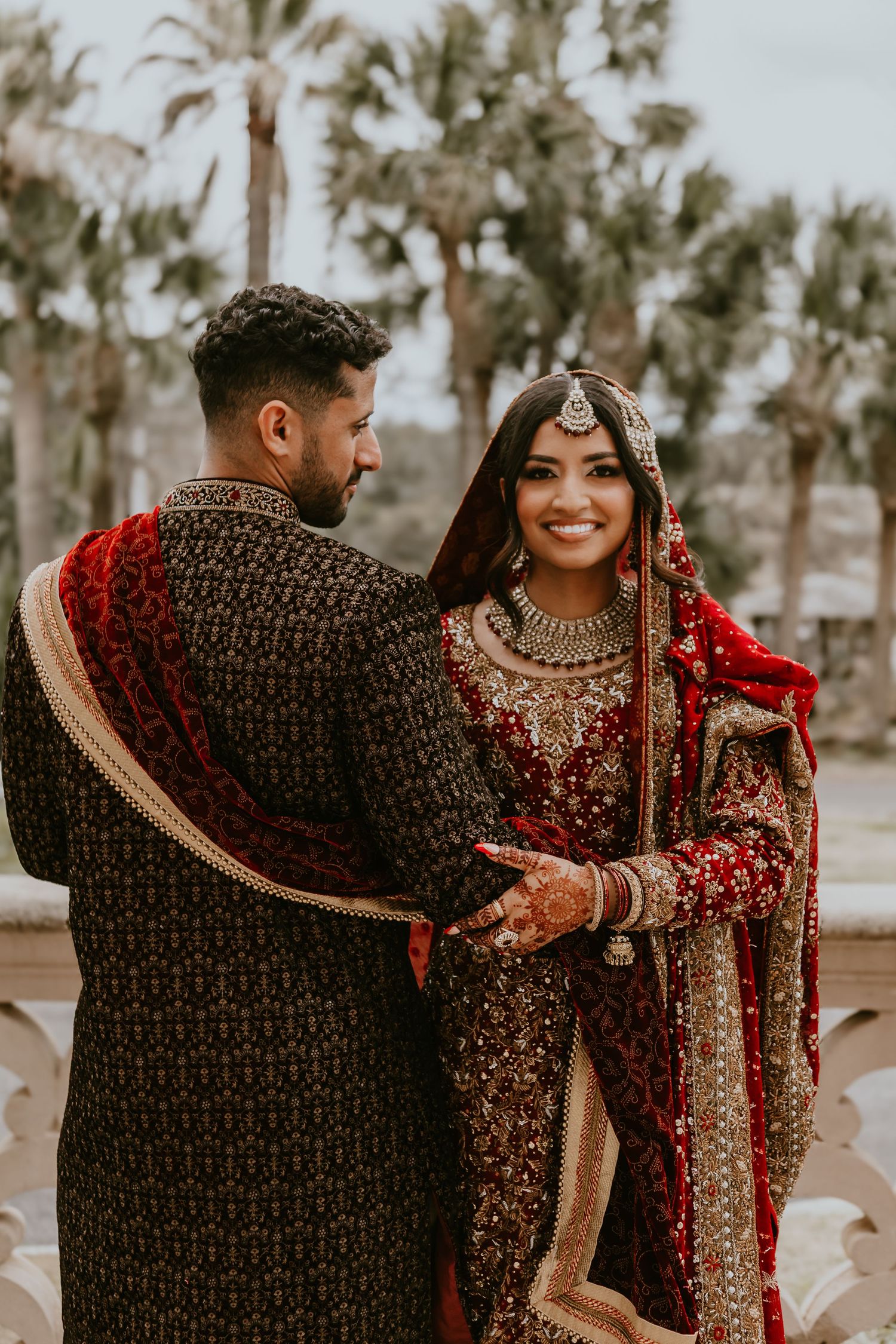 A couple in traditional South Asian wedding attire embrace outdoors, with the bride wearing a red and gold lehenga.
