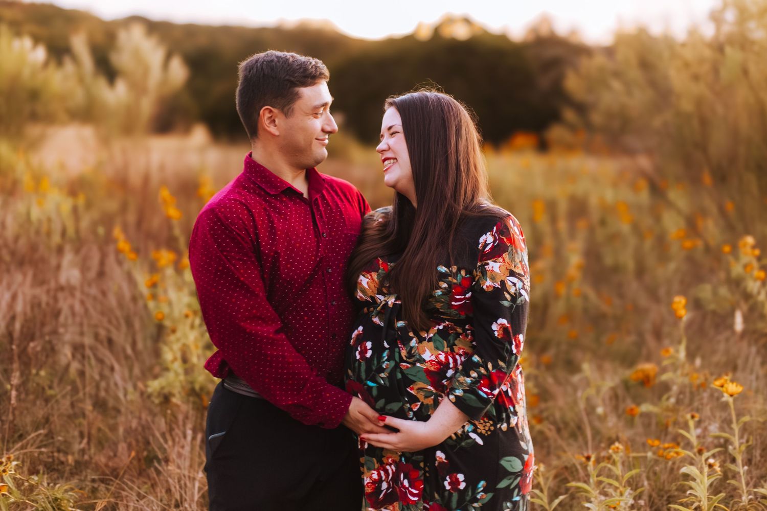 A couple in coordinating red and floral outfits embraces while smiling at each other in a golden autumn field at sunset.