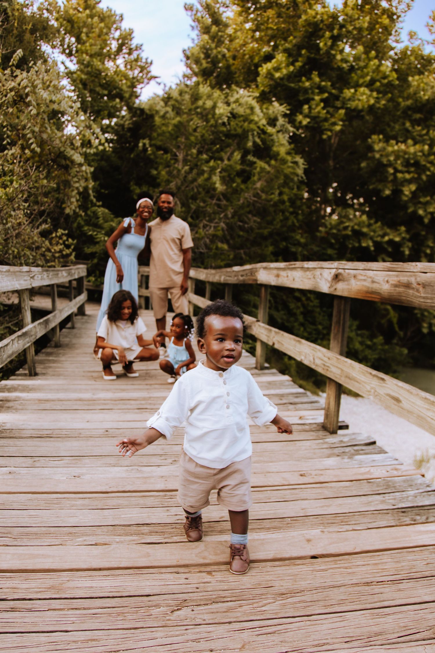Happy child in white shirt and khaki pants runs playfully on wooden boardwalk through nature while others walk behind.