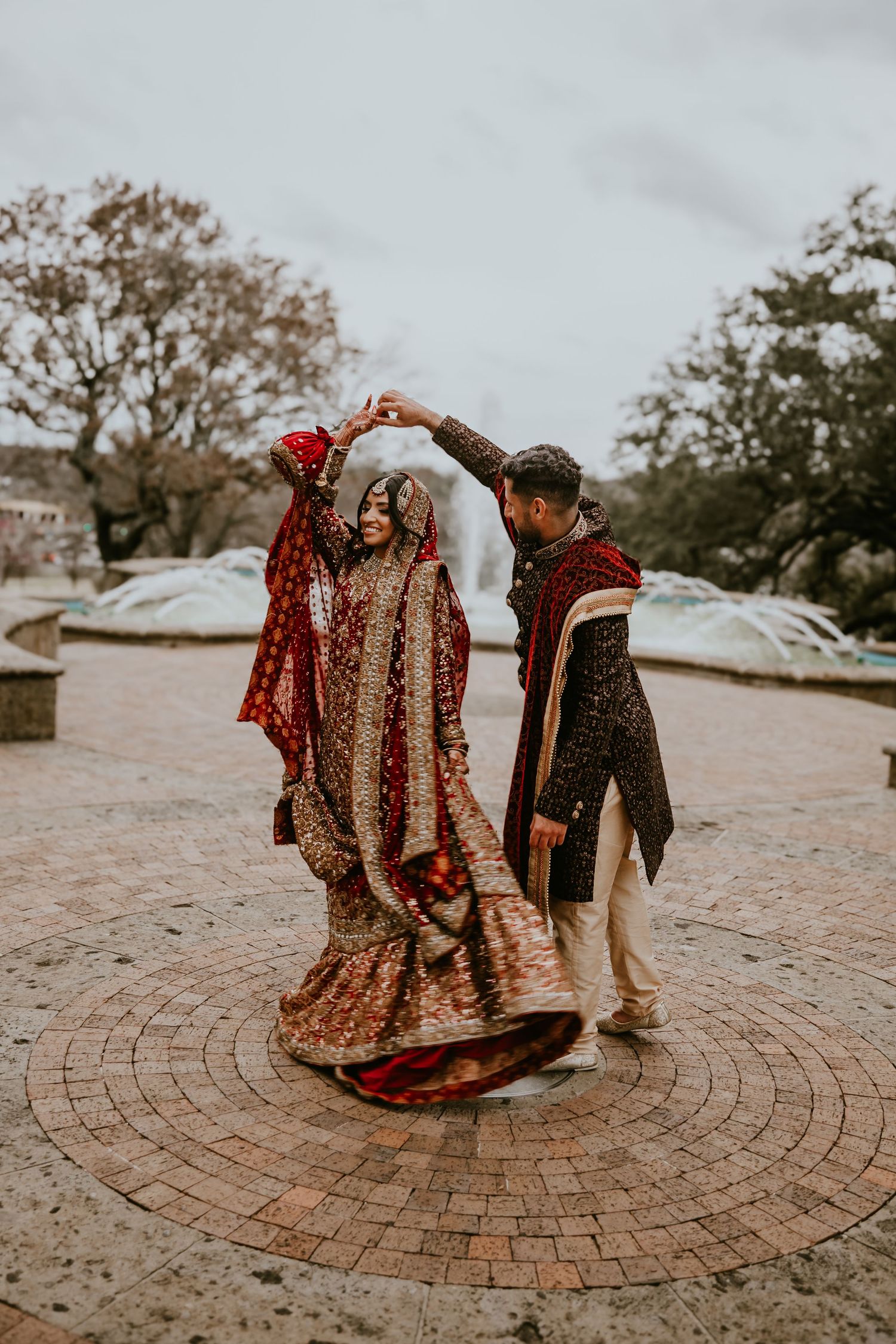 Couple in traditional South Asian wedding attire twirls on circular brick patio during intimate outdoor ceremony.